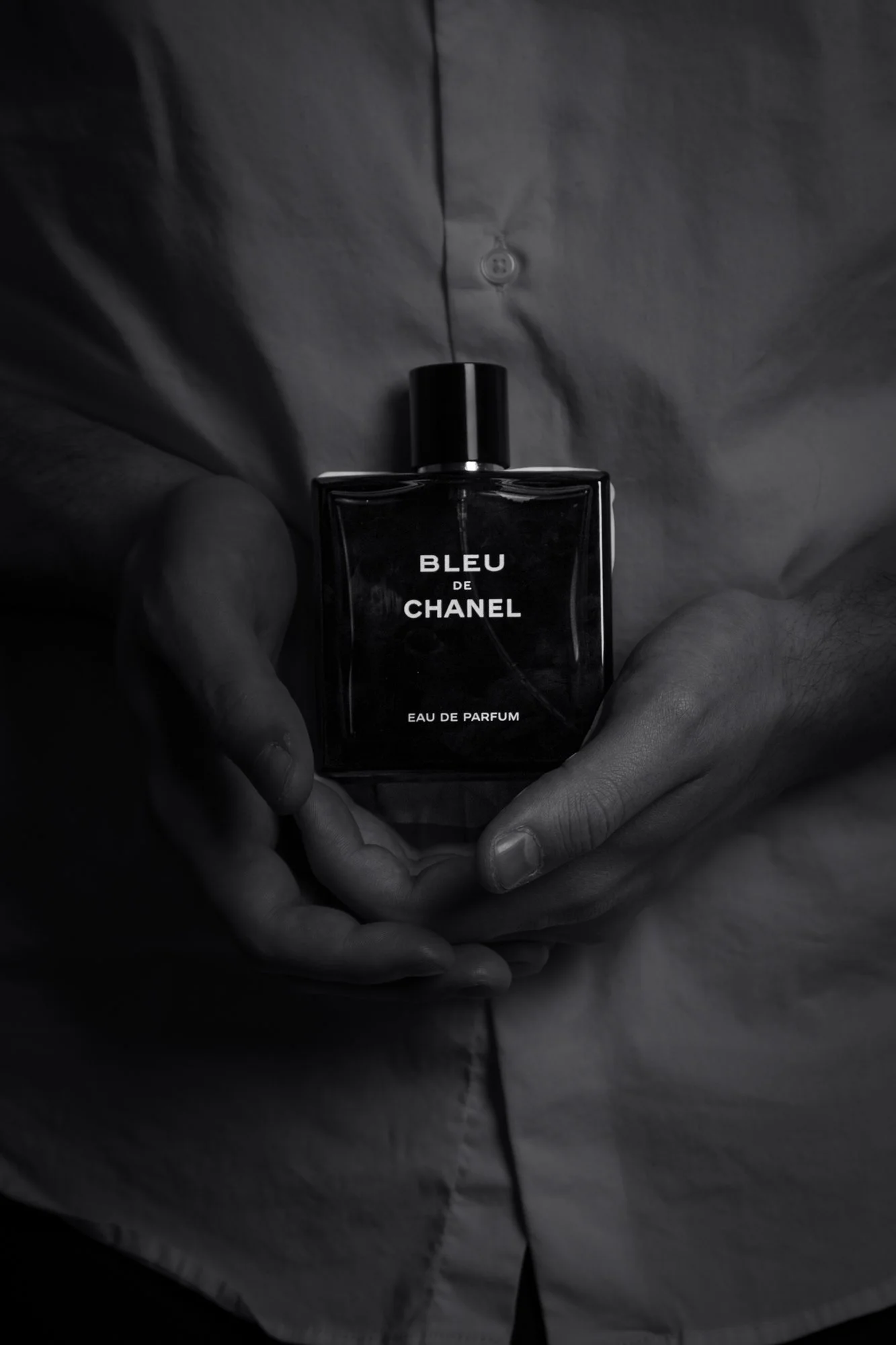 Close-up of a person holding a bottle of Bleu de Chanel Eau de Parfum against a dark background, with the person's torso and hands visible.