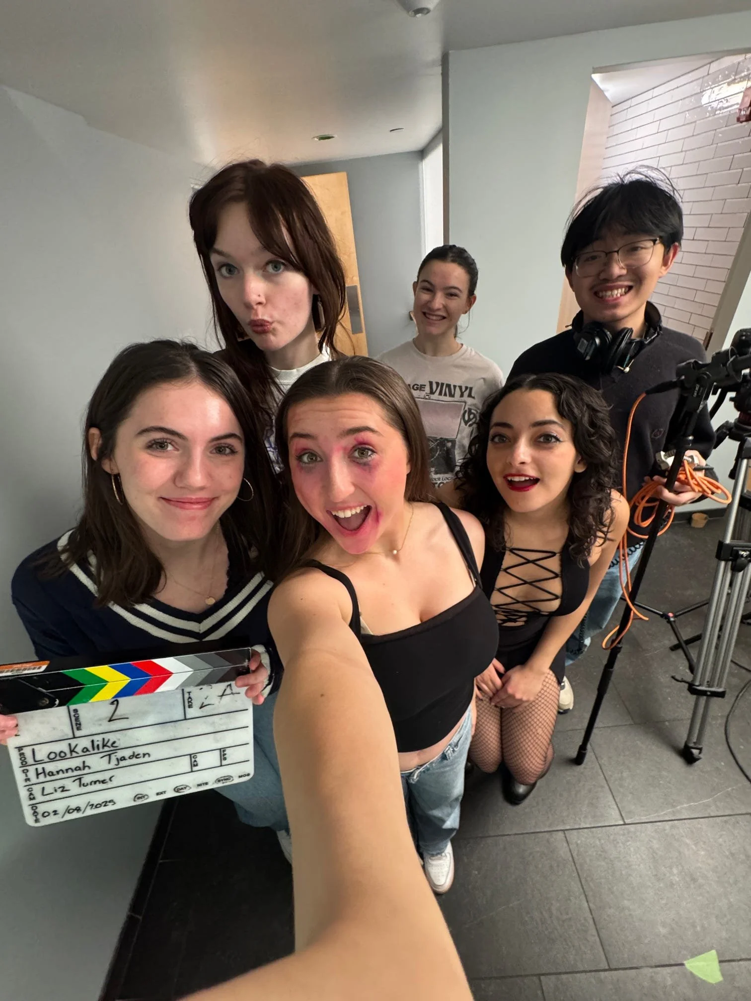 Group of six young women posing for a photo in an indoor setting, with filming equipment and a clapperboard in the foreground.