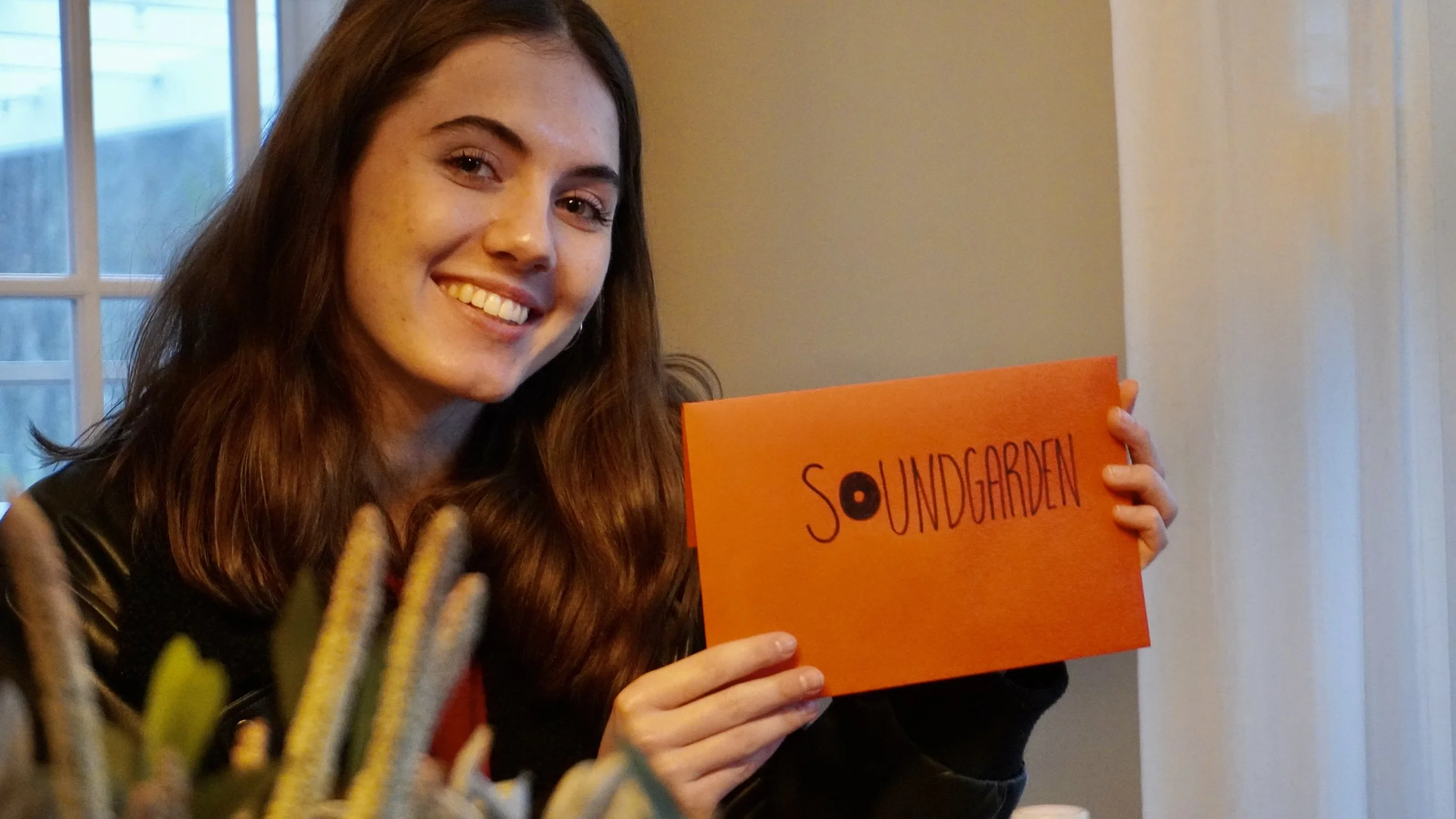 A young woman smiling and holding an orange sign with black writing, standing in a room with a window and a curtain in the background.