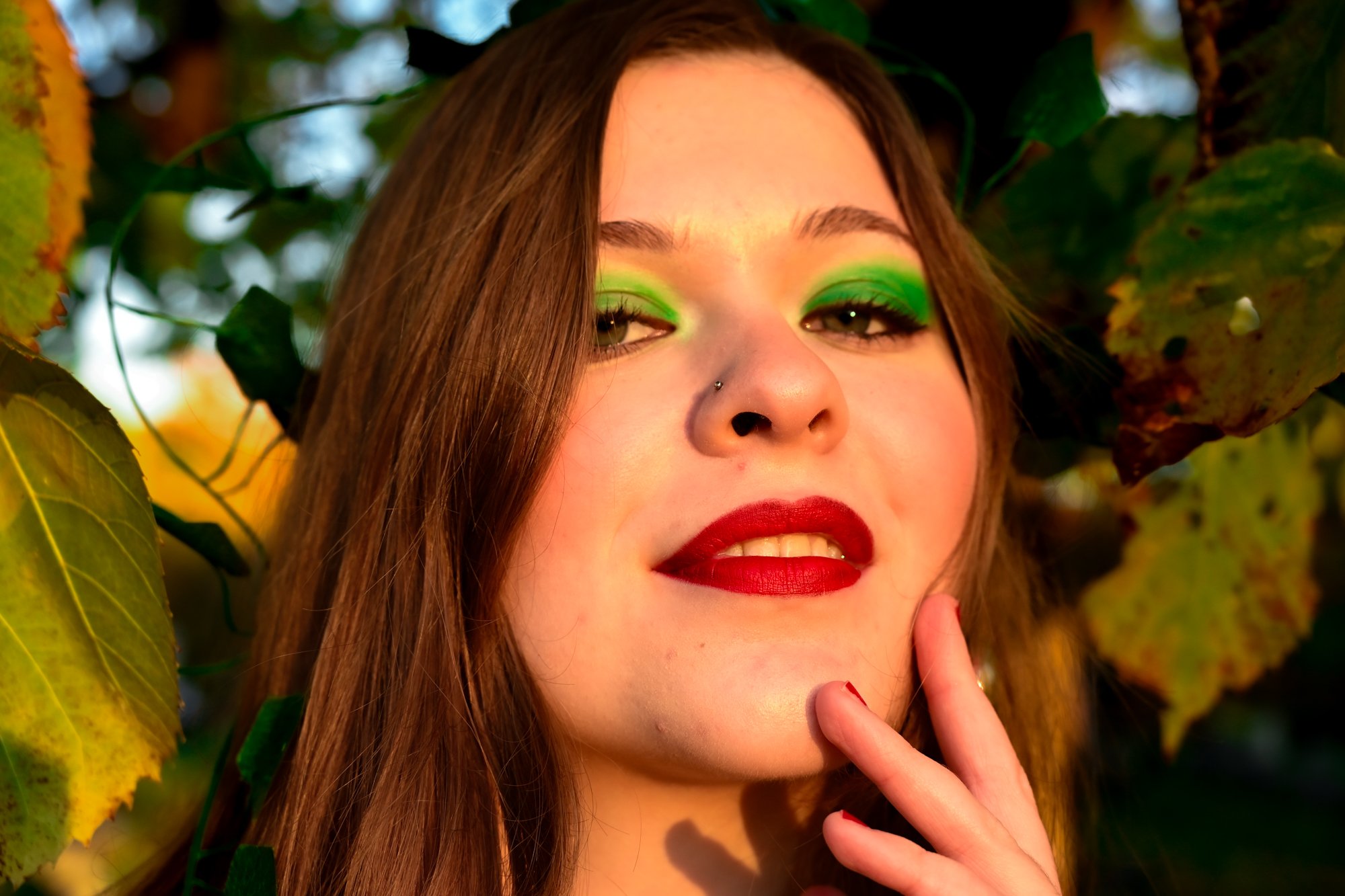 Close-up of a young woman with long brown hair, green eye makeup, red lipstick, and a nose piercing, surrounded by green and yellow leaves.