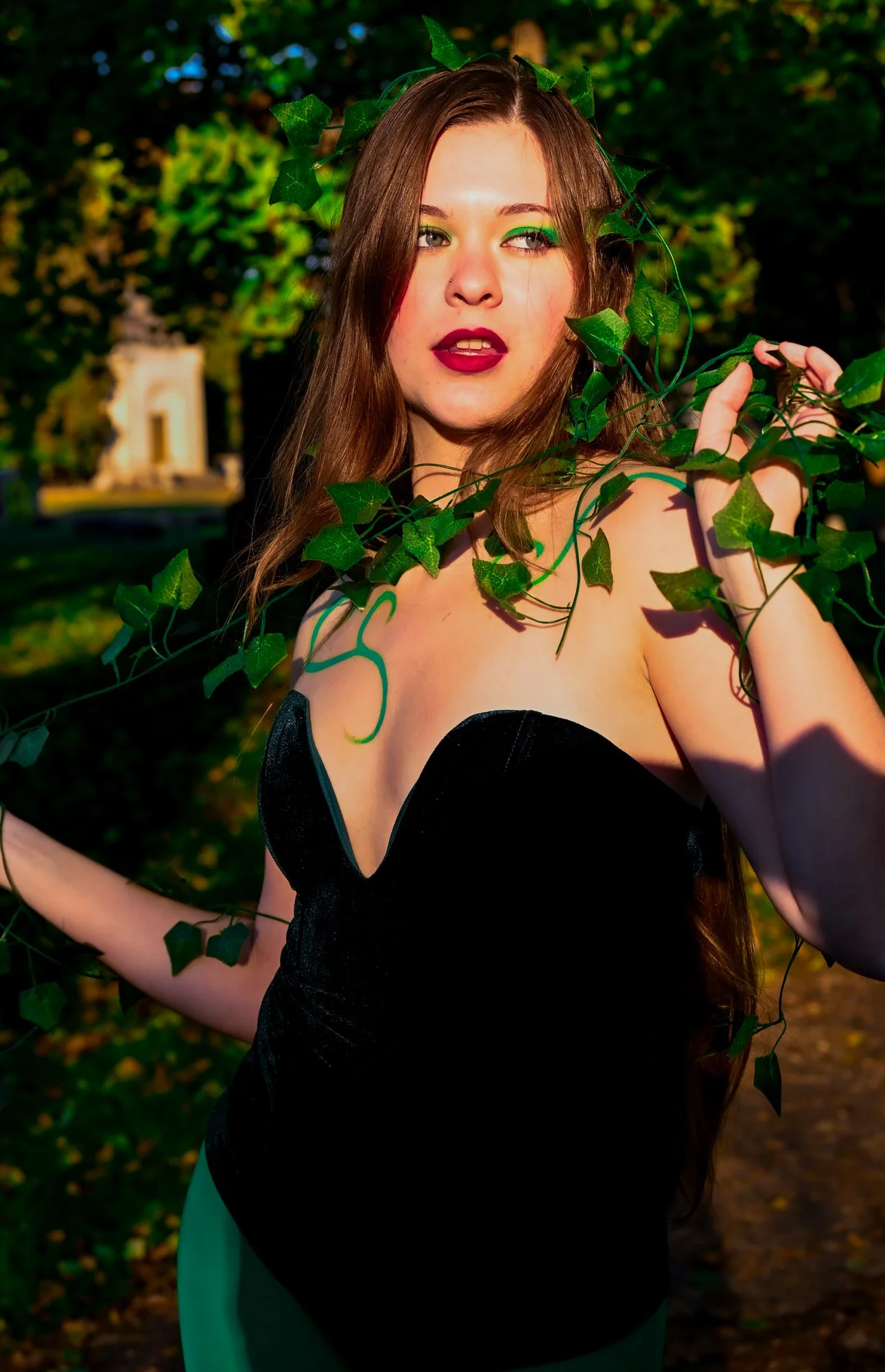 A young woman with long brown hair and makeup, wearing a black dress, is outdoors among green trees and leaves, with a blurred structure in the background, holding a vine with green leaves.