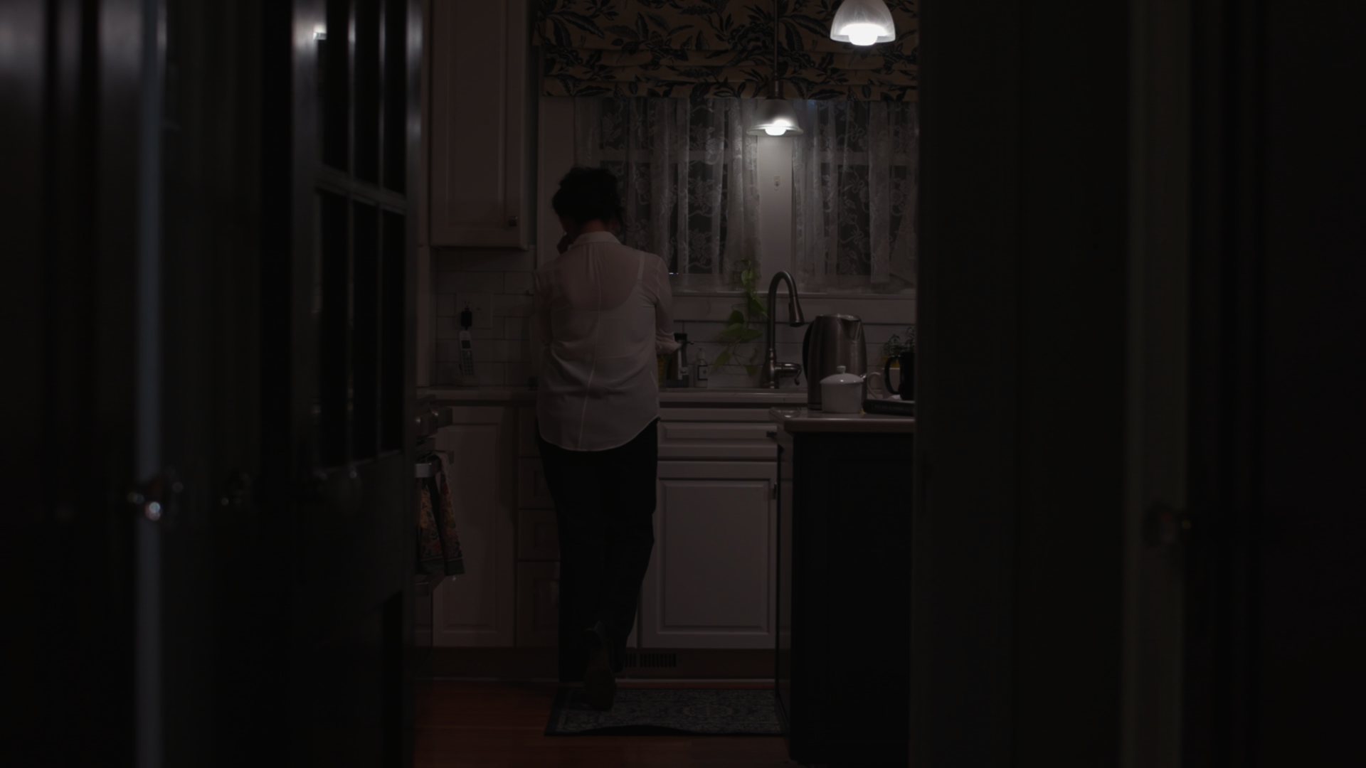 A person standing in a kitchen, viewed through a partially open door, with the person facing the sink area and talking on a phone.