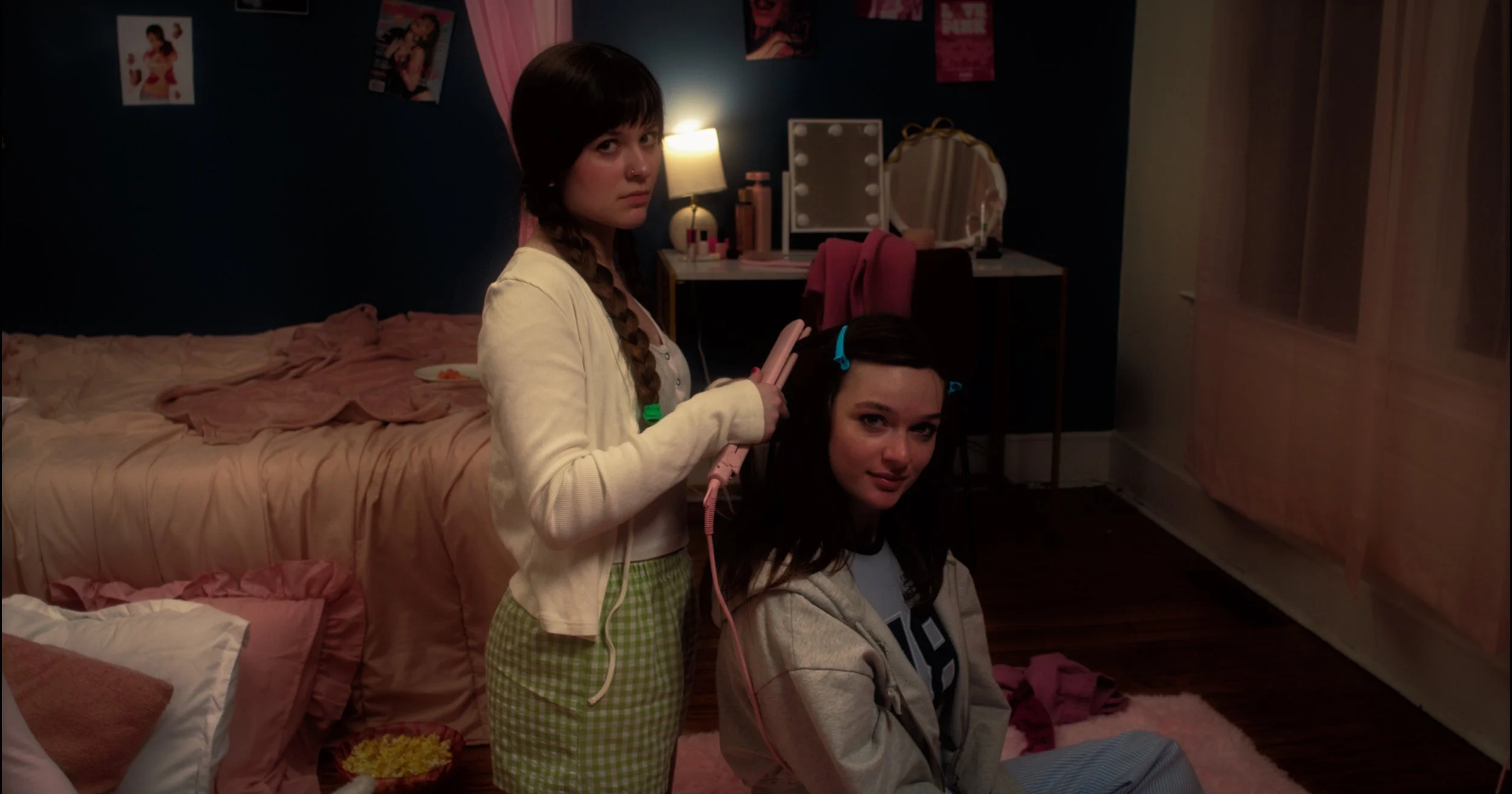 Two young women in a dimly lit bedroom, one standing using a curling iron, and the other sitting on the floor, both looking at the camera. The room has pink and dark-colored walls, a bed with pink bedding, a vanity mirror with light bulbs, and various photos and posters on the wall.