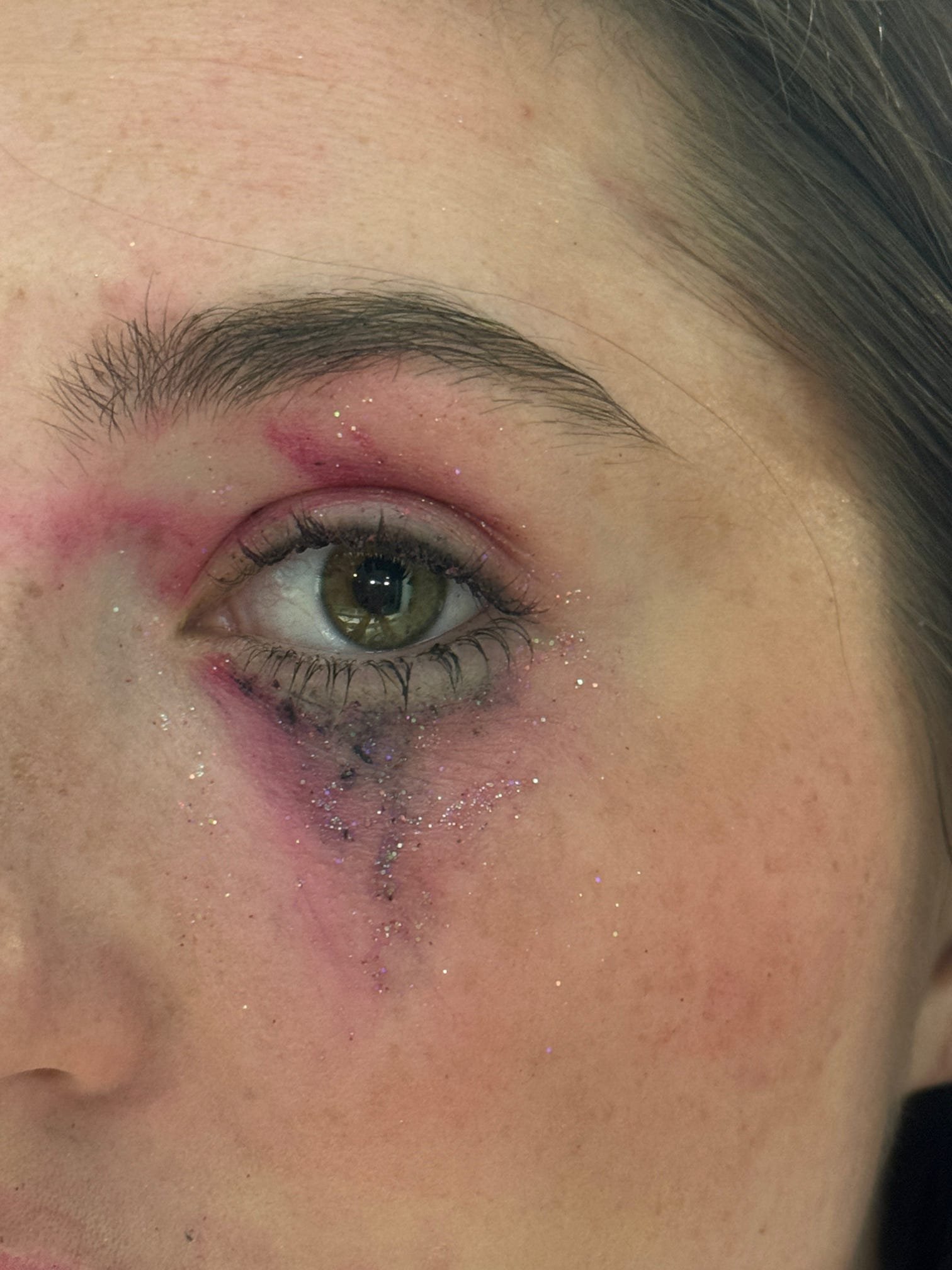 Close-up of a woman's eye with makeup, sparkles, and glitter, showing her eyebrow, eyelid, and part of her cheek and nose.