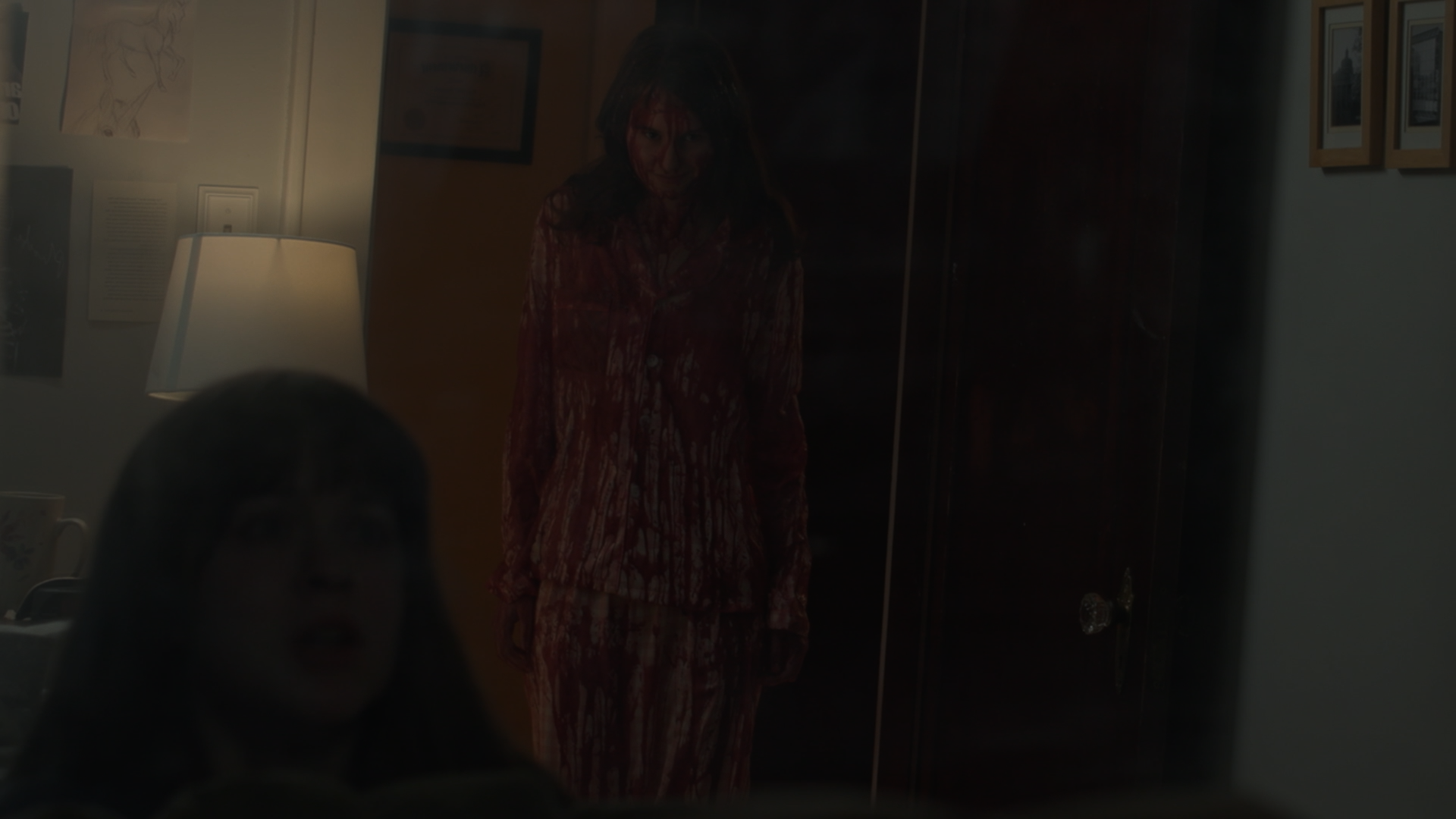 A woman in a bloody costume standing behind a glass door, with another person's head partially visible in the foreground.