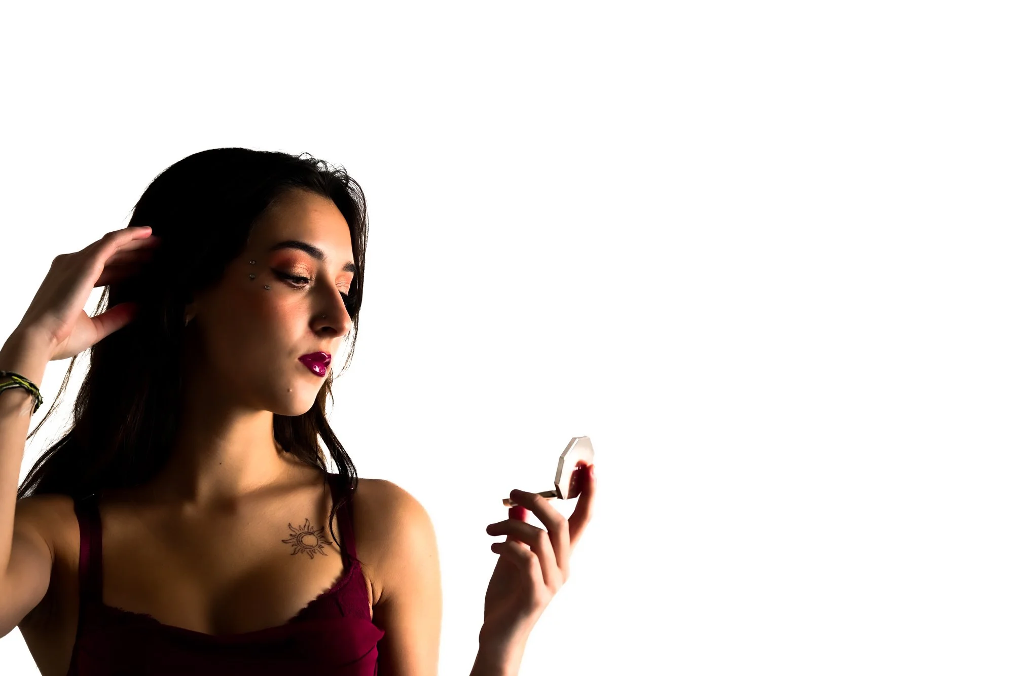A young woman with dark hair, makeup, and tattoos, wearing a burgundy top, is looking at her phone and touching her hair.