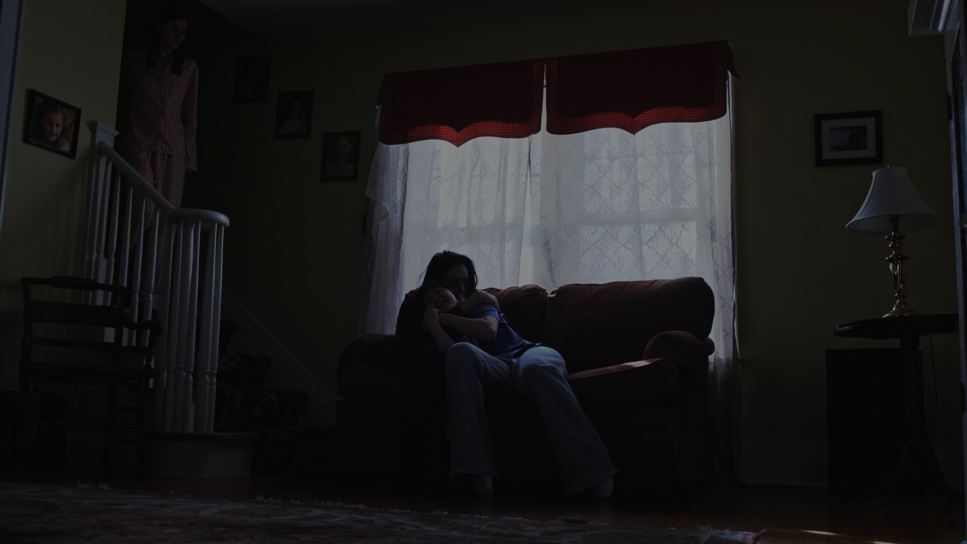 A woman and a child sit on a sofa in a dimly lit living room, with sunlight filtering through lace curtains behind them. Another woman stands on the staircase to the left, and framed photographs decorate the walls.