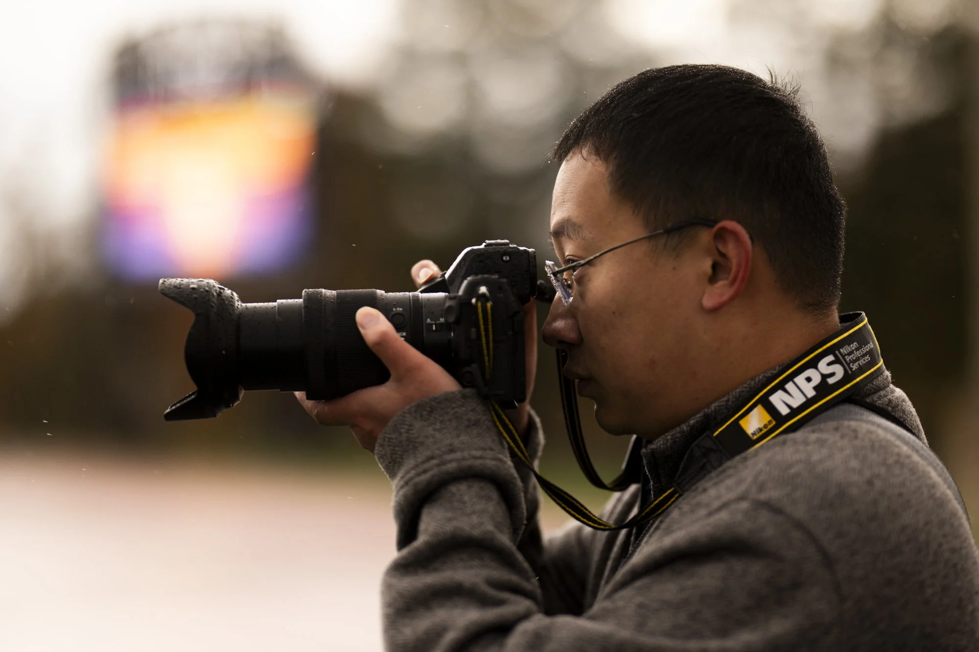 A man with glasses and short dark hair taking a photograph with a professional camera. He is holding the camera up to his eye and wearing a dark hoodie and a Nikon strap around his neck. The background is blurred.