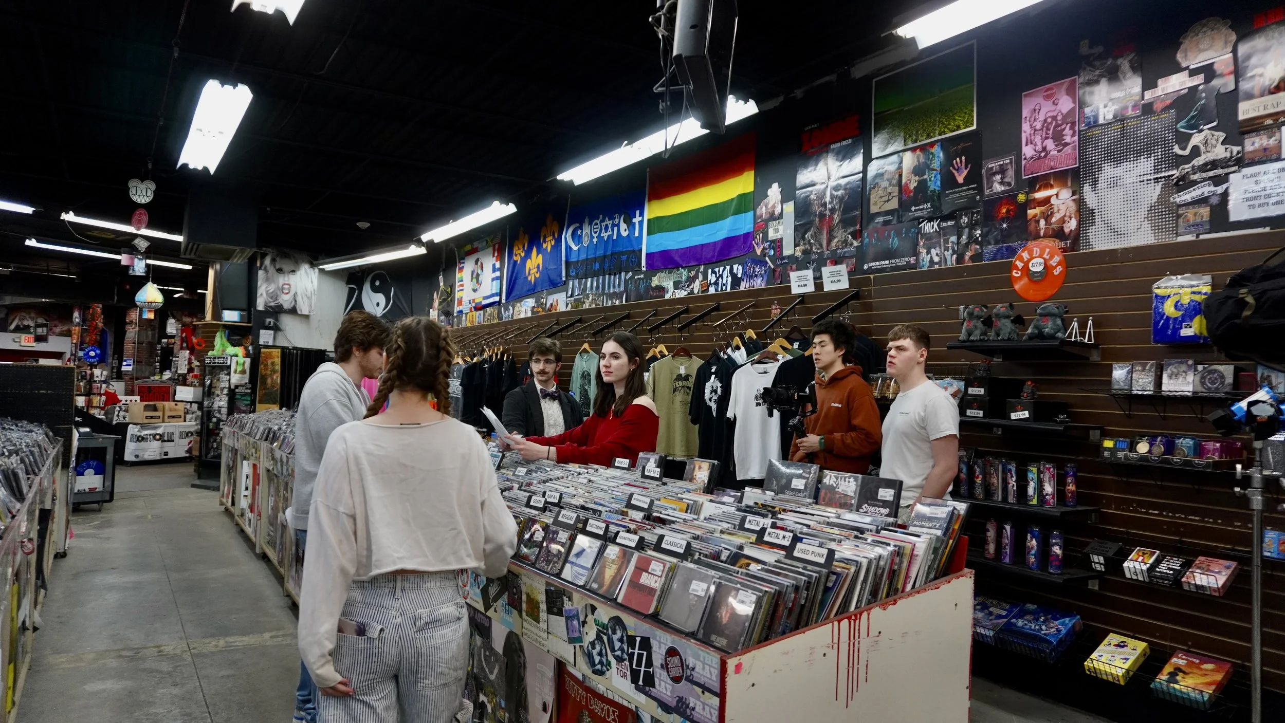 People shopping at a record store with posters and flags on the wall, including a rainbow pride flag and flags with fleur-de-lis symbols, selling vinyl records and merchandise.