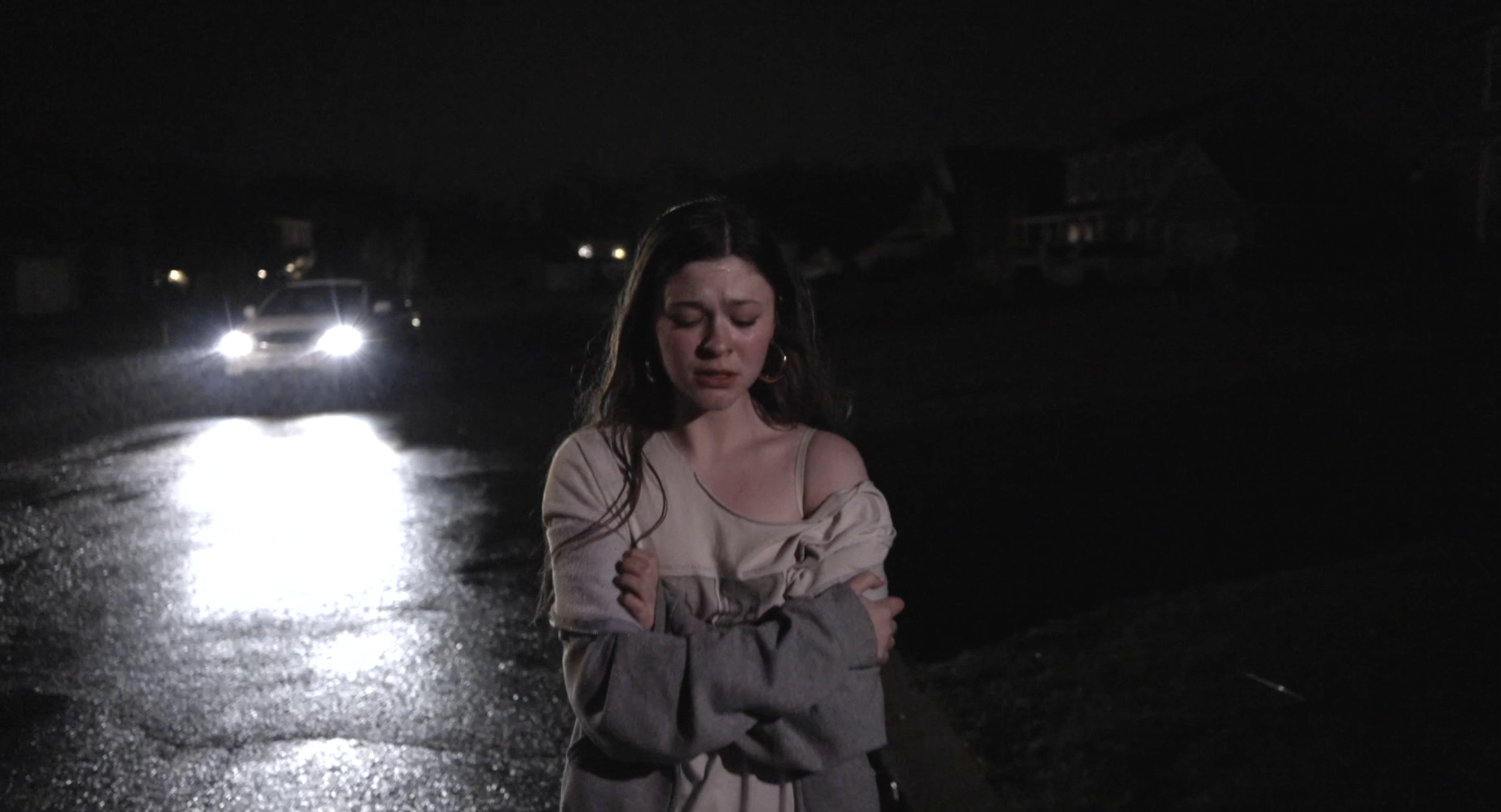 A woman with long dark hair stands on a wet dark street at night, illuminated by headlights in the background. She appears distressed with her eyes closed and arms crossed over her chest.