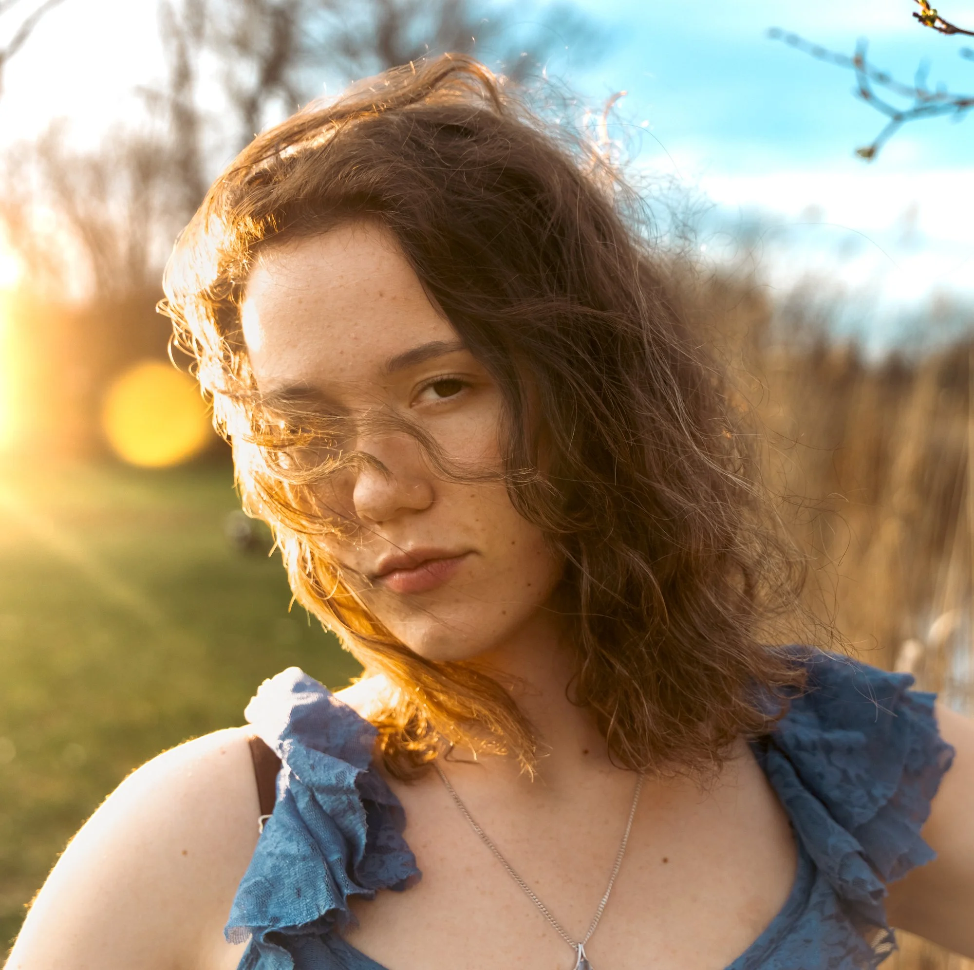 A woman with wavy, reddish-brown hair and freckles, wearing a blue and black-patterned top, stands outdoors in natural sunlight with blurred trees and sky in the background.