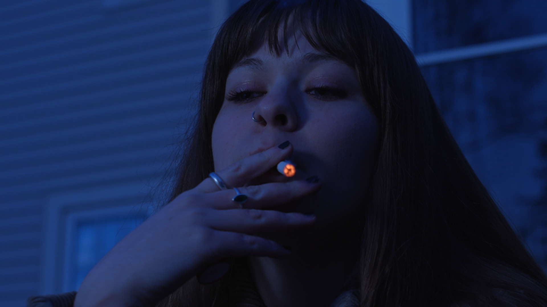 A young woman with brown hair and a nose ring is smoking a cigarette at night, illuminated by a blue light.