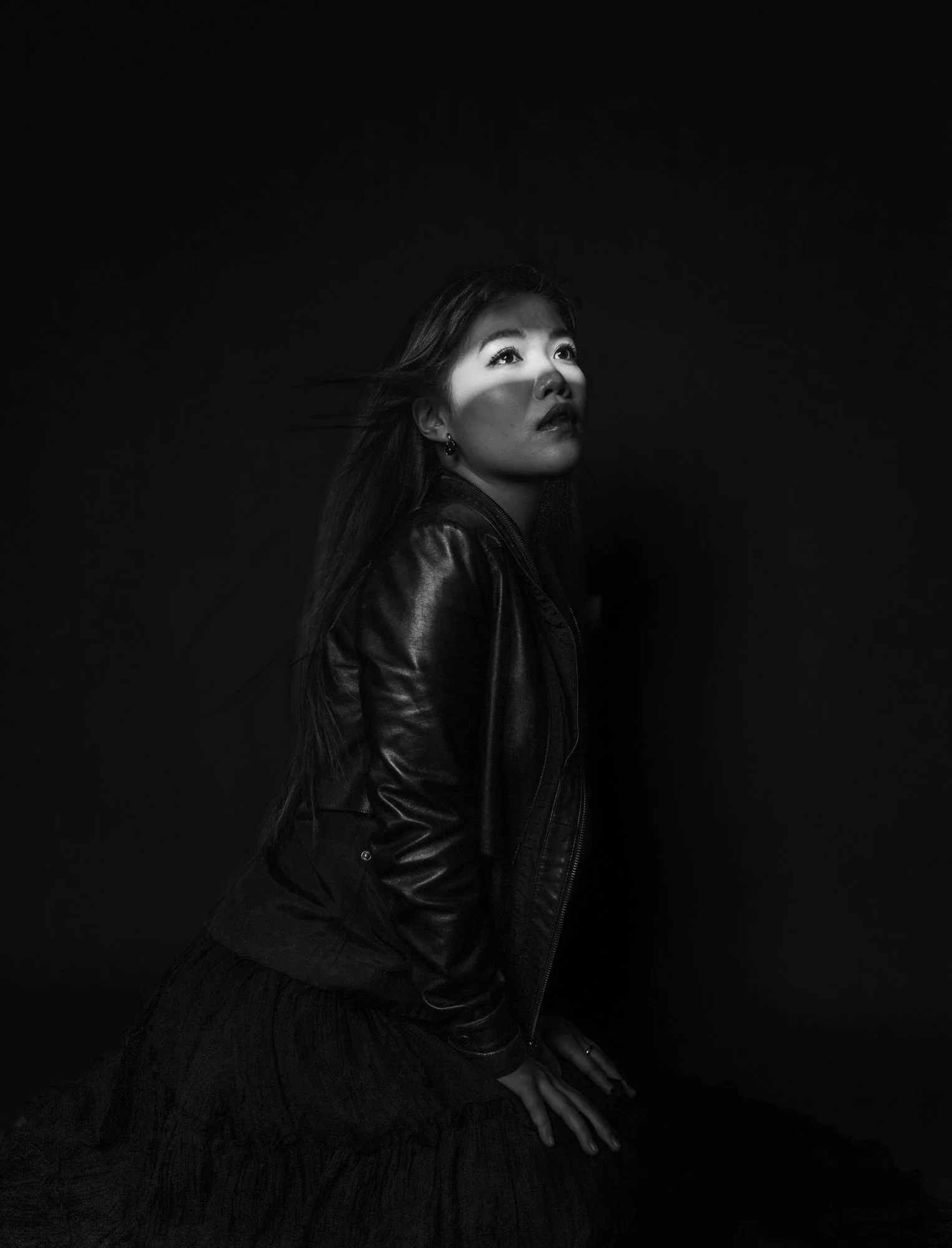 Black and white photo of a woman with long hair, looking upward with a soft expression, wearing a leather jacket, standing in front of a dark background with a beam of light hitting her face.