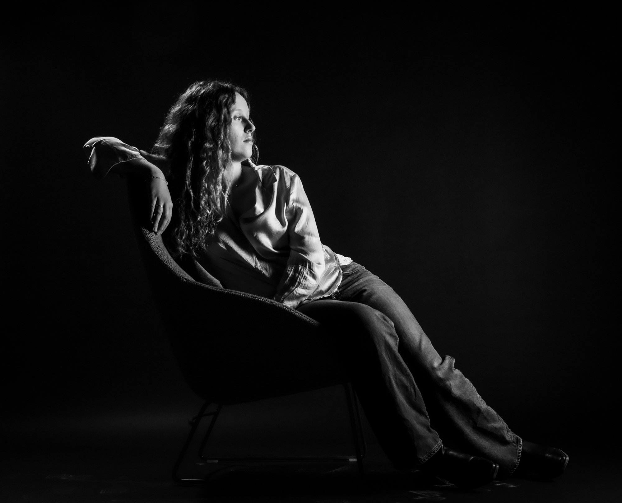 A black and white photo of a woman with long, curly hair sitting sideways in a chair against a black background, looking to the right with a serious expression, wearing a loose, light-colored shirt and jeans.