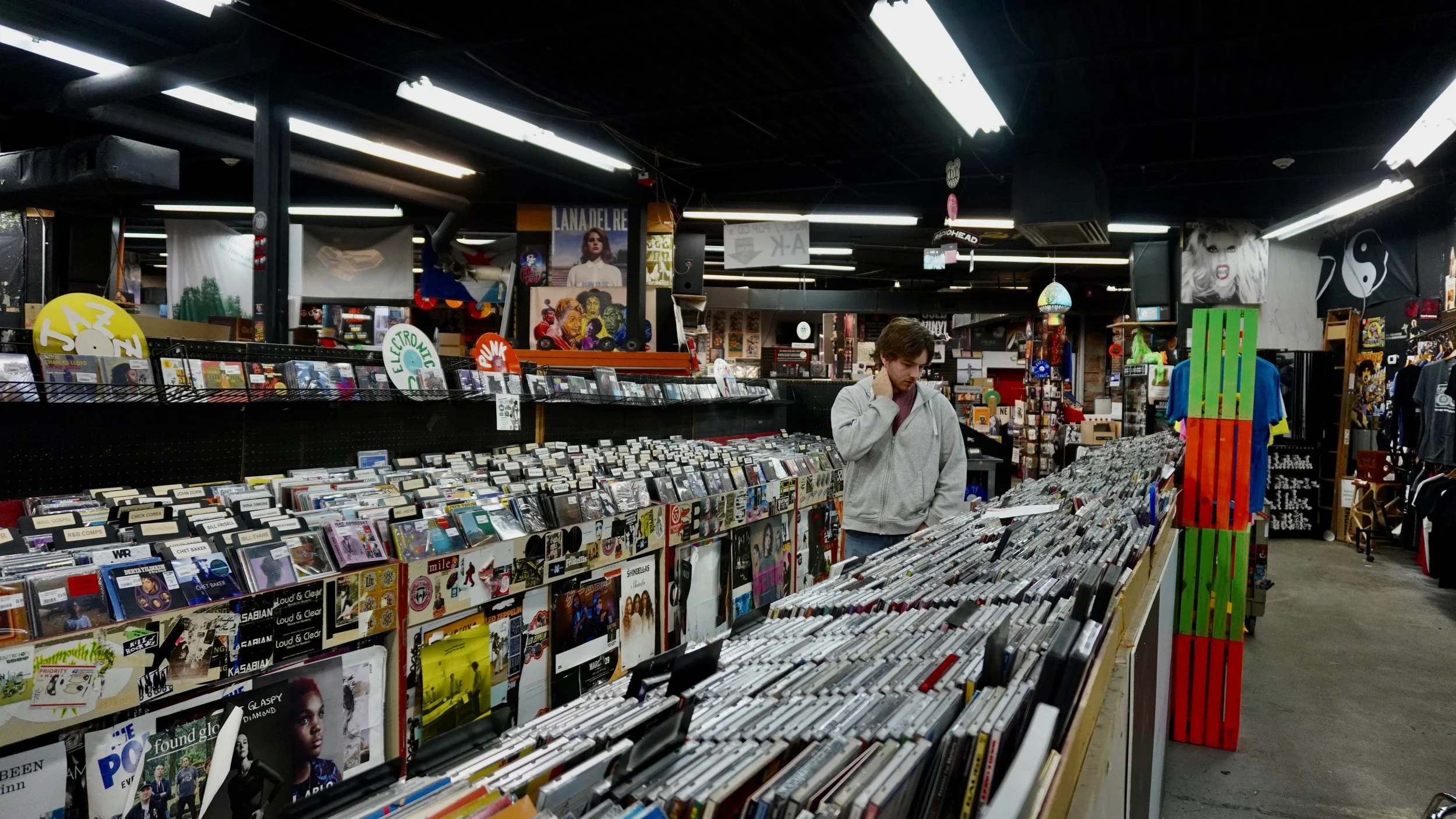 Inside a record store with rows of vinyl records, posters, and artwork, including a woman with blonde hair on a poster, and a man in a gray hoodie browsing the records.