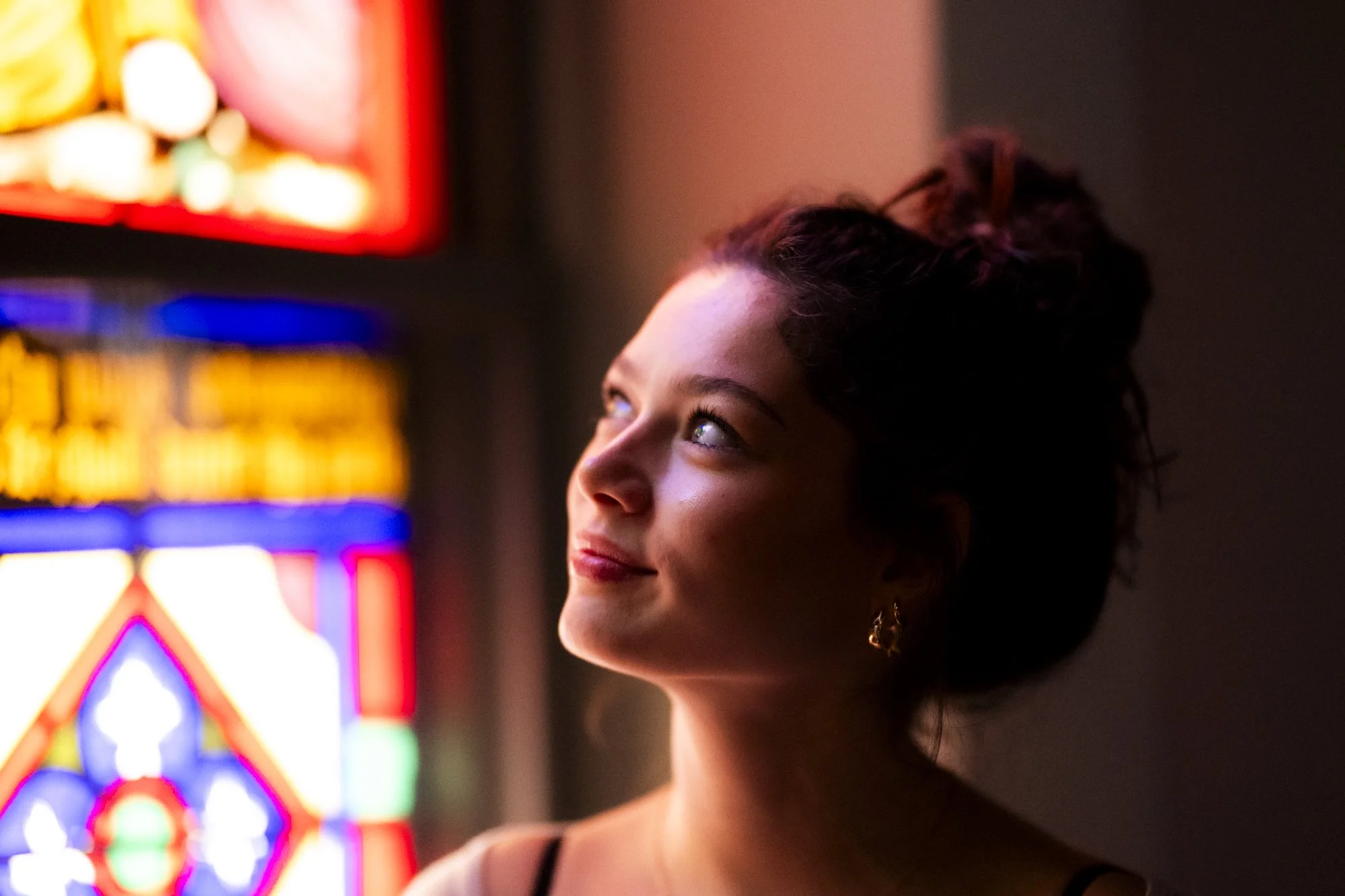 A young woman with brown hair styled in a bun, light makeup, and earrings, looking at colorful stained glass lit up background in a dimly lit setting.