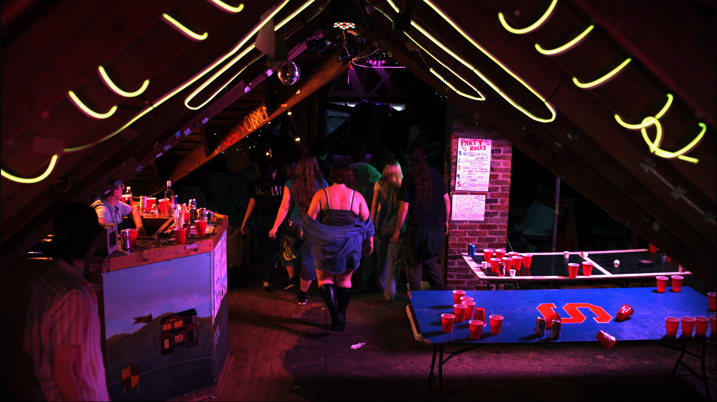 People playing beer pong at a backyard party in a dimly lit attic with neon lights and a disco ball.