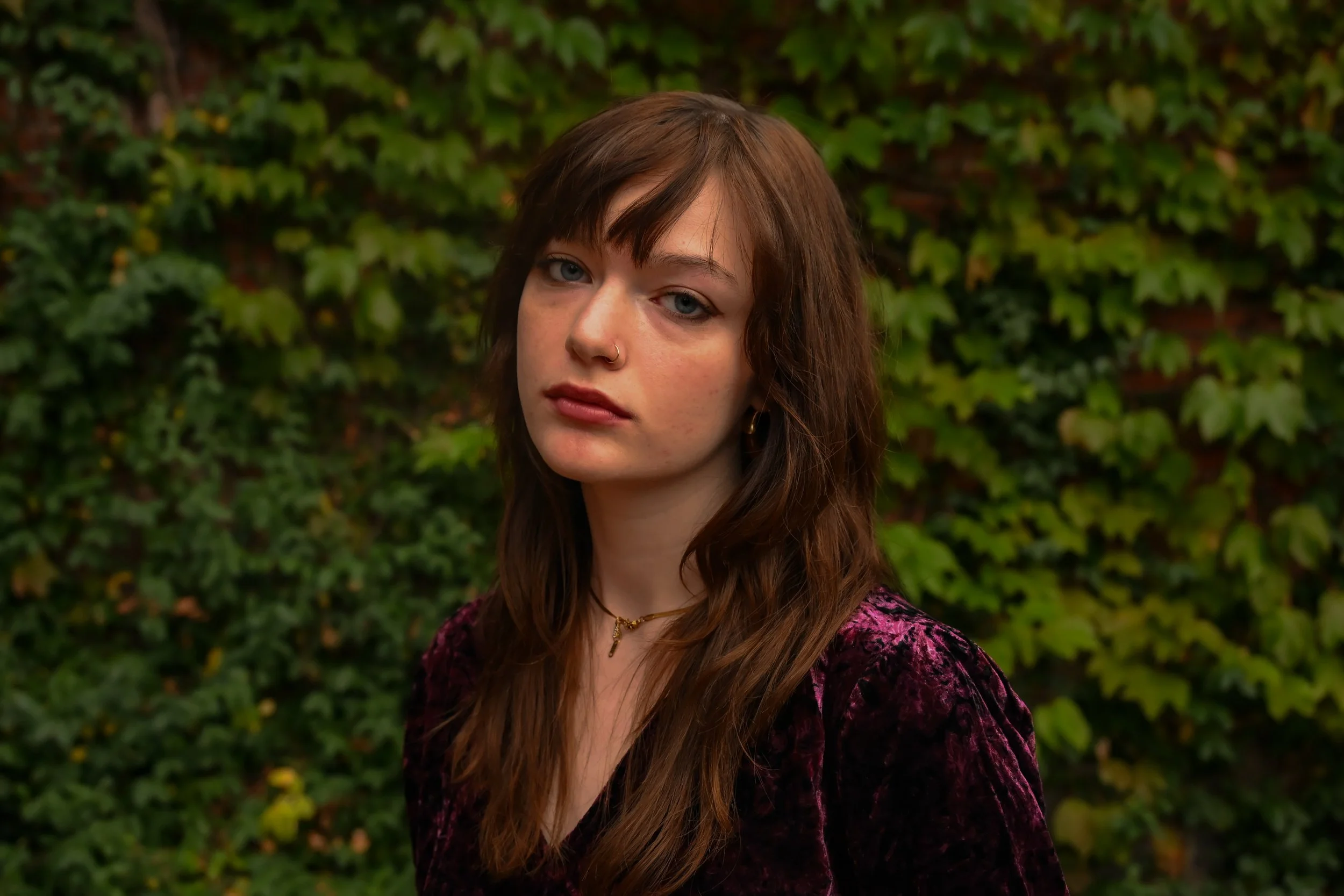 A young woman with long brown hair and light skin is standing outdoors in front of a green leafy hedge. She is wearing a purple velvet jacket and has a small nose piercing, with a neutral expression on her face.