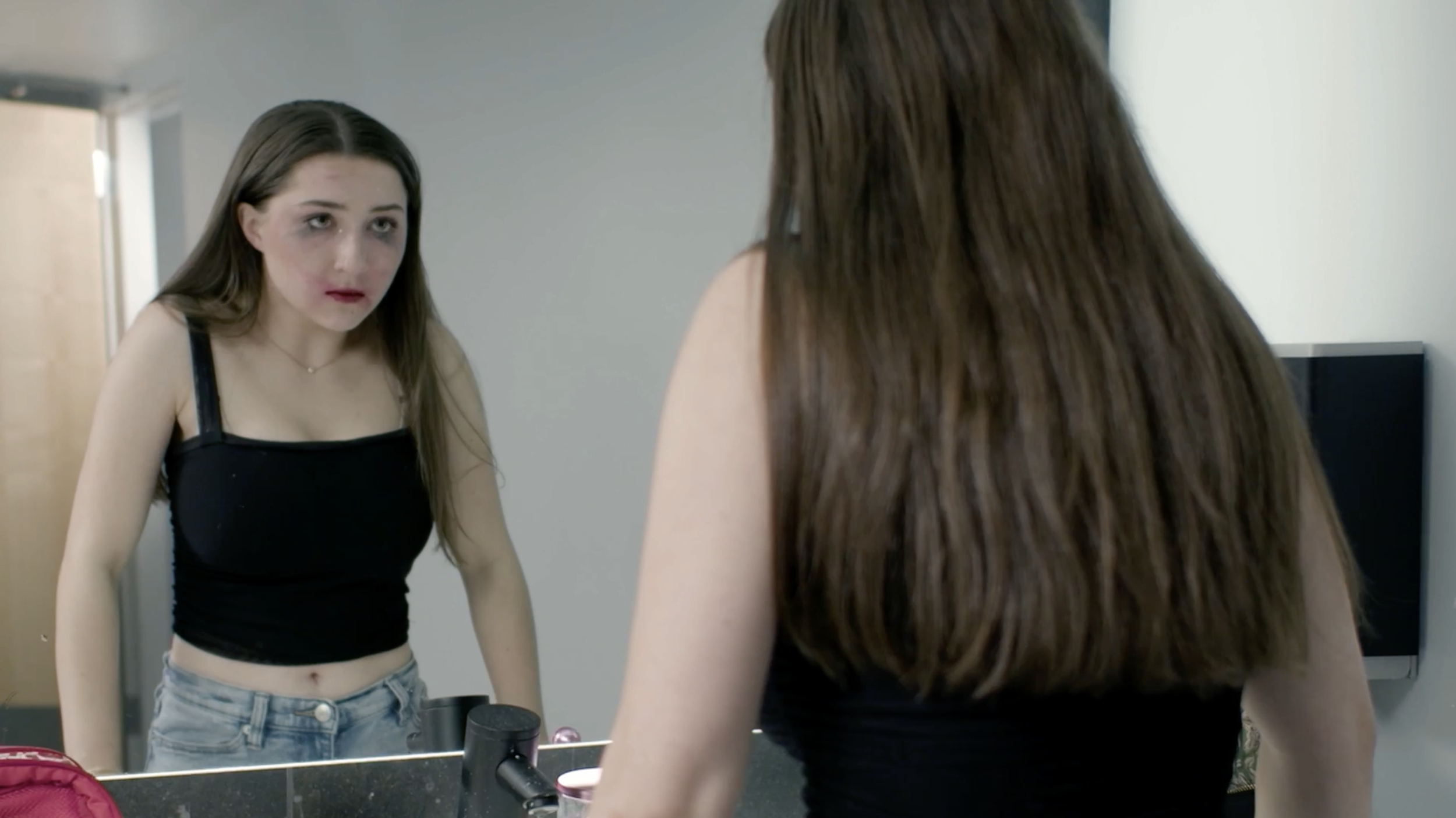 A young woman with bruises and dark circles under her eyes looking at herself in a bathroom mirror.