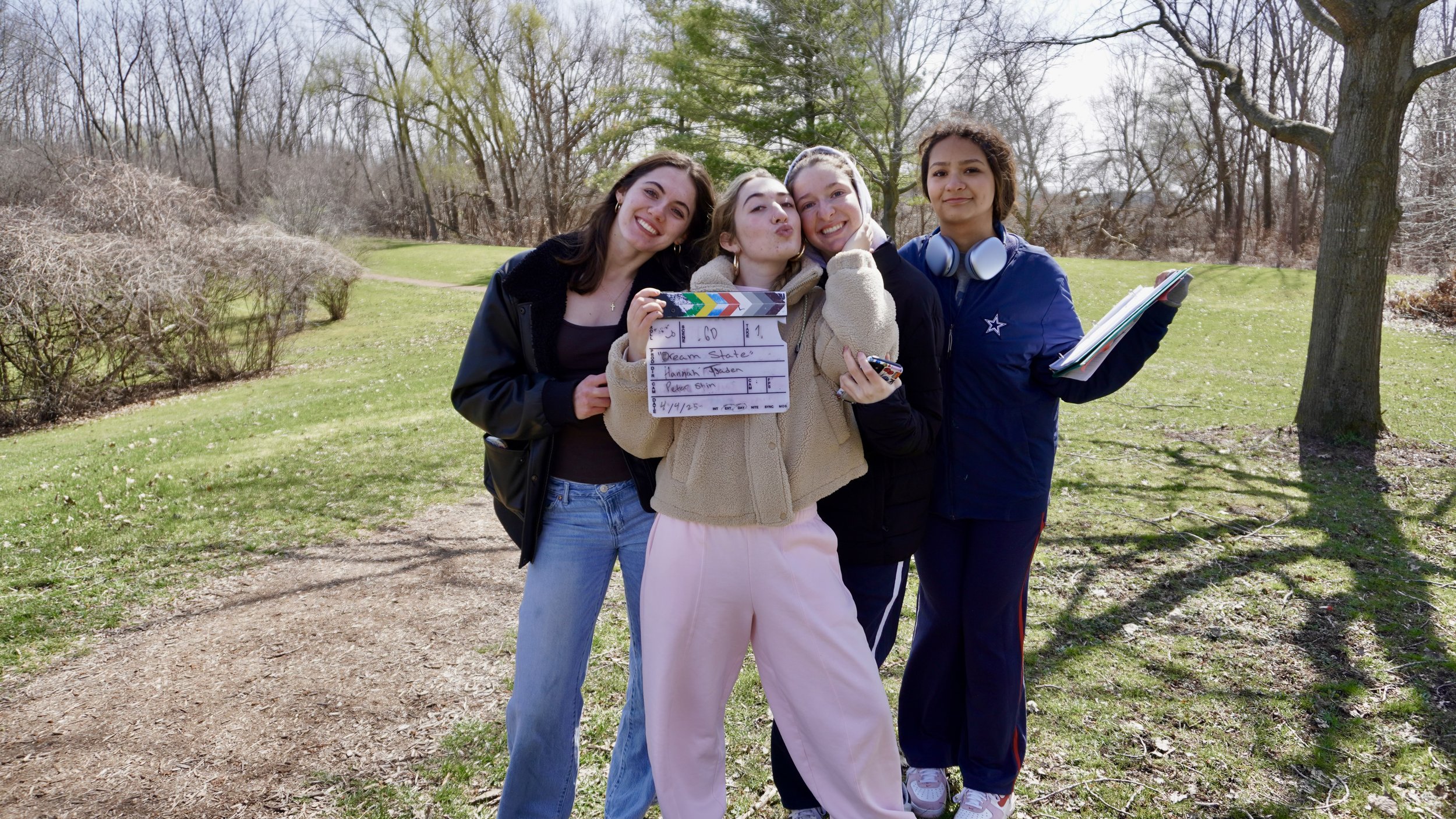 Four young women standing close together outdoors in a park, smiling at the camera, with trees and grass in the background. One of them is holding a clapperboard, and another is holding a stack of notebooks or folders.