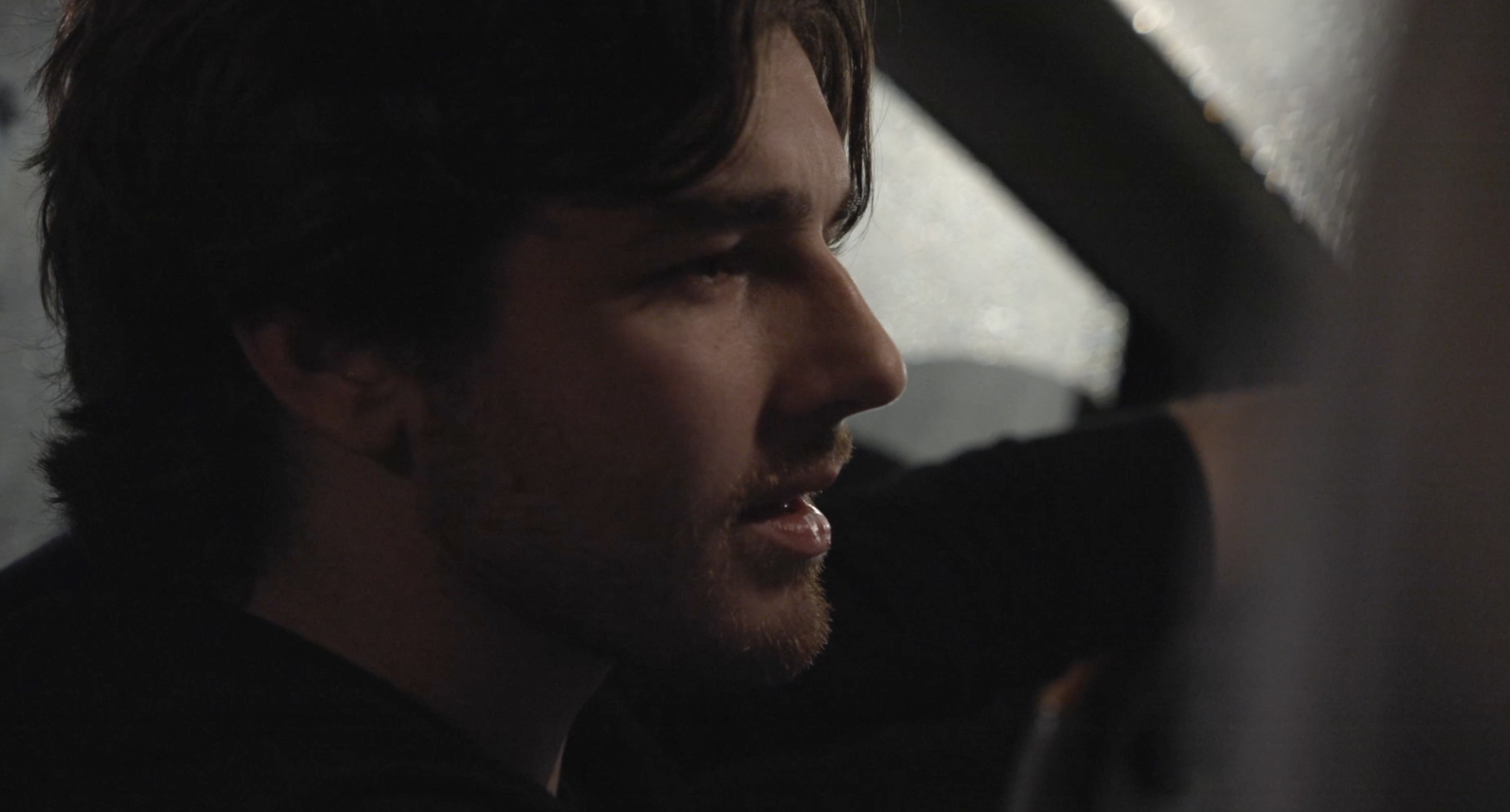 A man with dark hair and facial hair is looking downward, with his eyes closed or squinting, in a dimly lit setting.