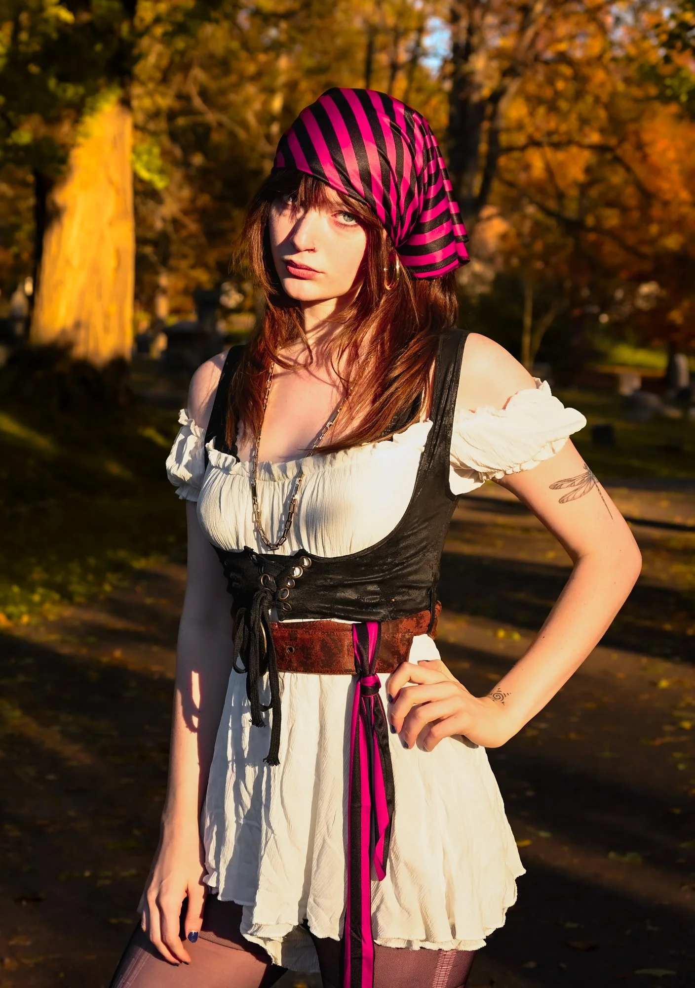 A woman with shoulder-length brown hair, blue eyes, and light skin posing outdoors during fall. She is wearing a pink and black striped headscarf, a white off-shoulder top with puffed sleeves, a black corset-style overlay, a brown belt with pink acce