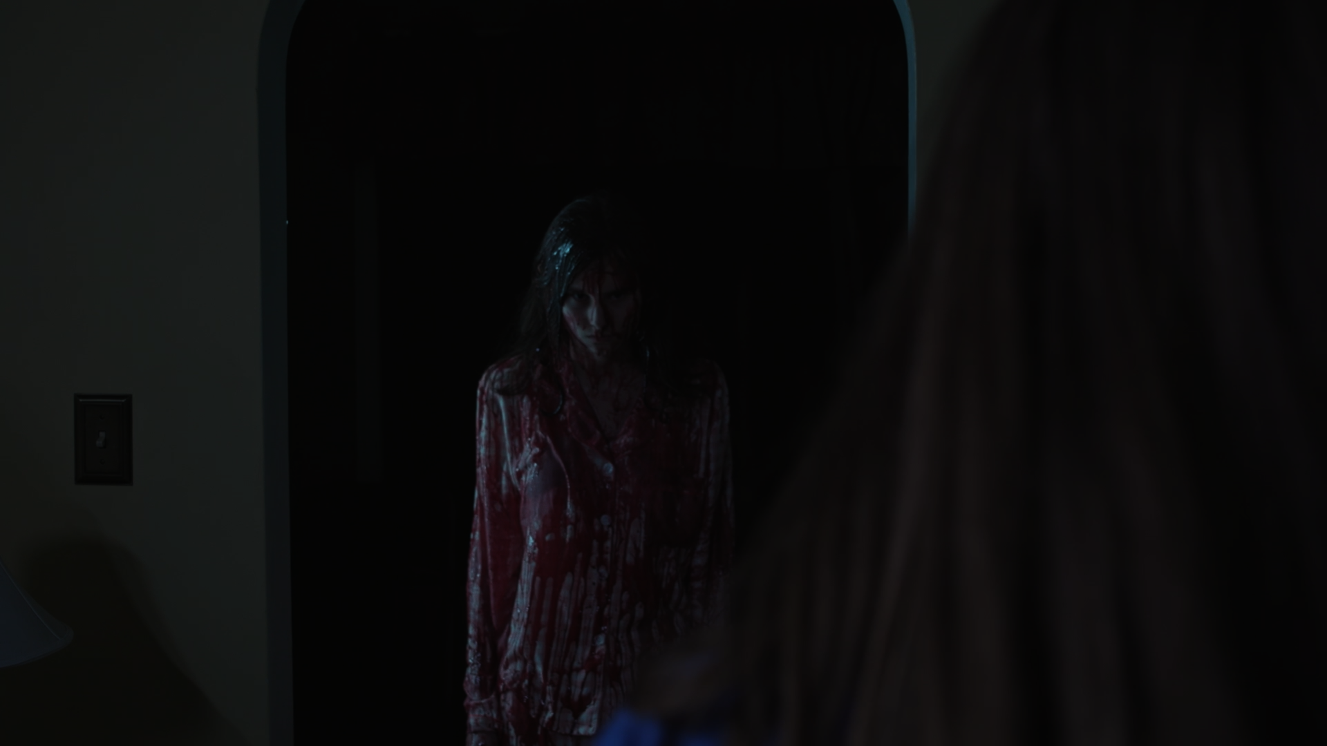 A woman with long hair looking at her reflection in a mirror, wearing a bloodstained shirt in a dark room.