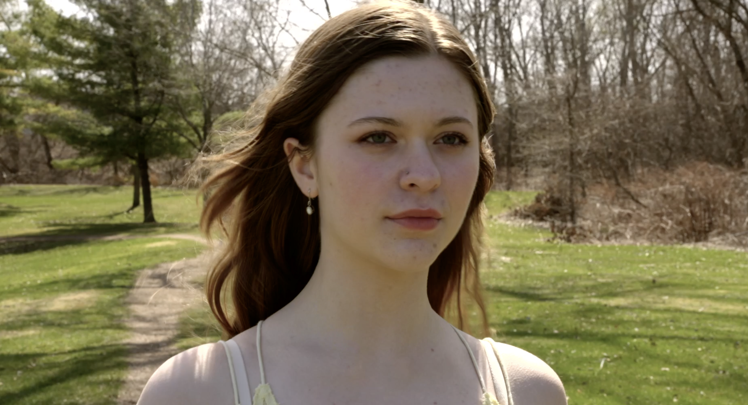 A young woman with long, reddish-brown hair standing outdoors in a park during daytime, with trees and grass in the background.
