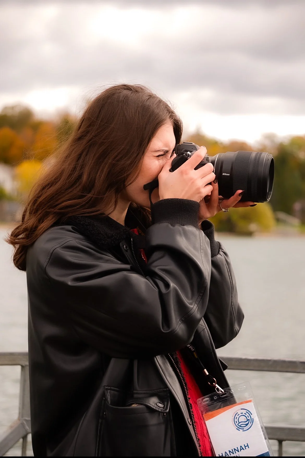A woman with brown hair, wearing a black jacket, takes a photo with a camera outdoors, with trees and cloudy sky in the background.