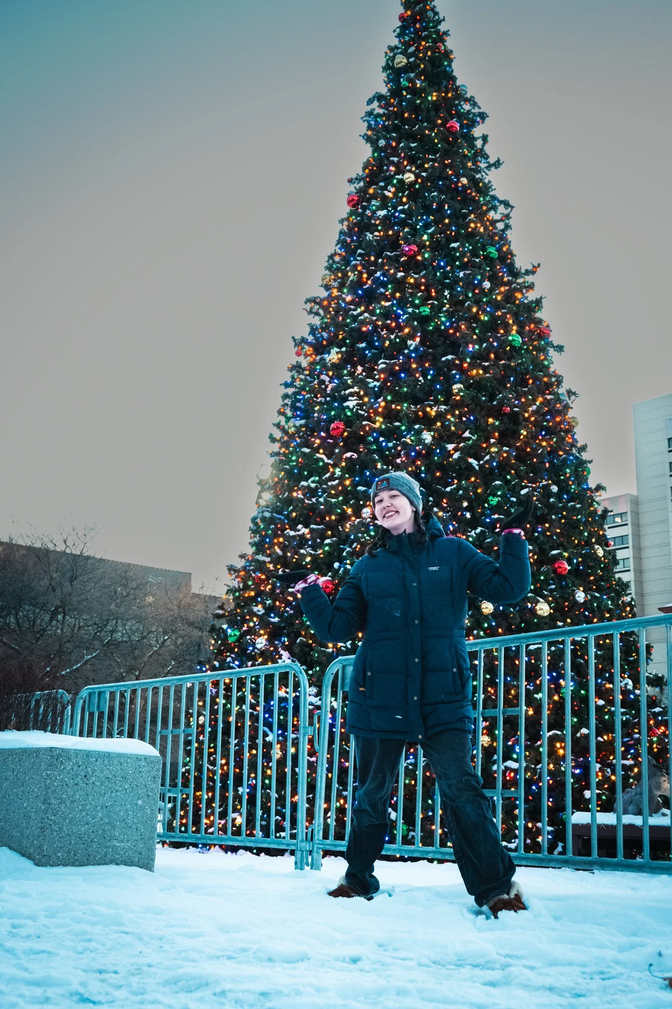 A person in a winter coat and beanie stands in front of a large, decorated Christmas tree outdoors, with snow on the ground and city buildings in the background.