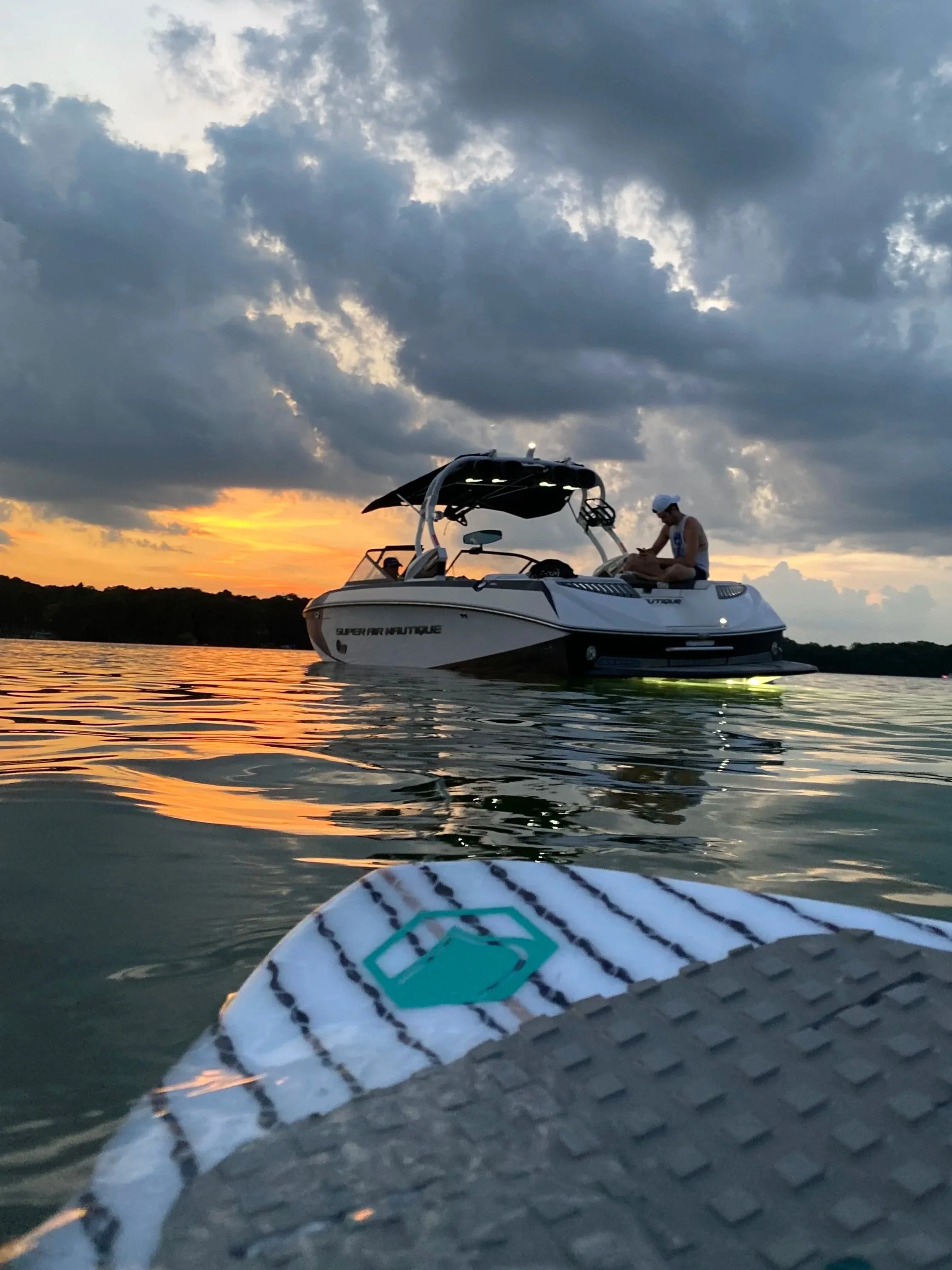 Close-up of a stand-up paddleboard with a logo, floating on water, with a boat and a person sitting on it in the background at sunset.