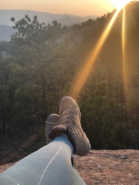 Person wearing hiking boots and jeans sitting on a rock, with legs crossed, overlooking a forested landscape at sunset with sunlight streaming down.