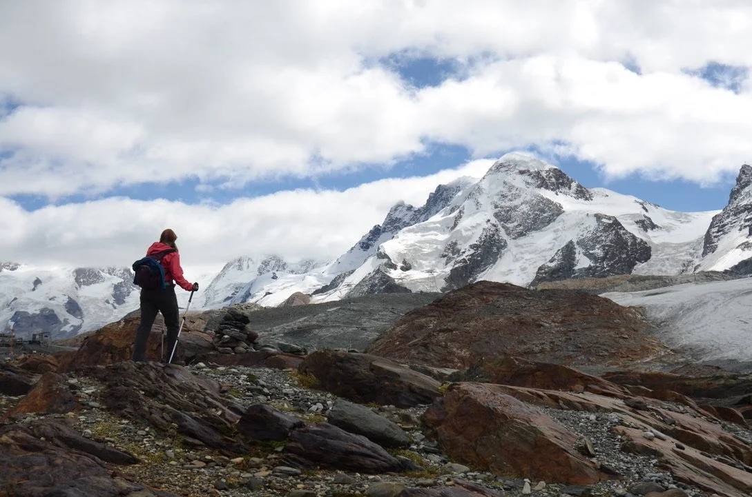 A hiker in a red jacket walking on rocky terrain with snow-capped mountains in the background.