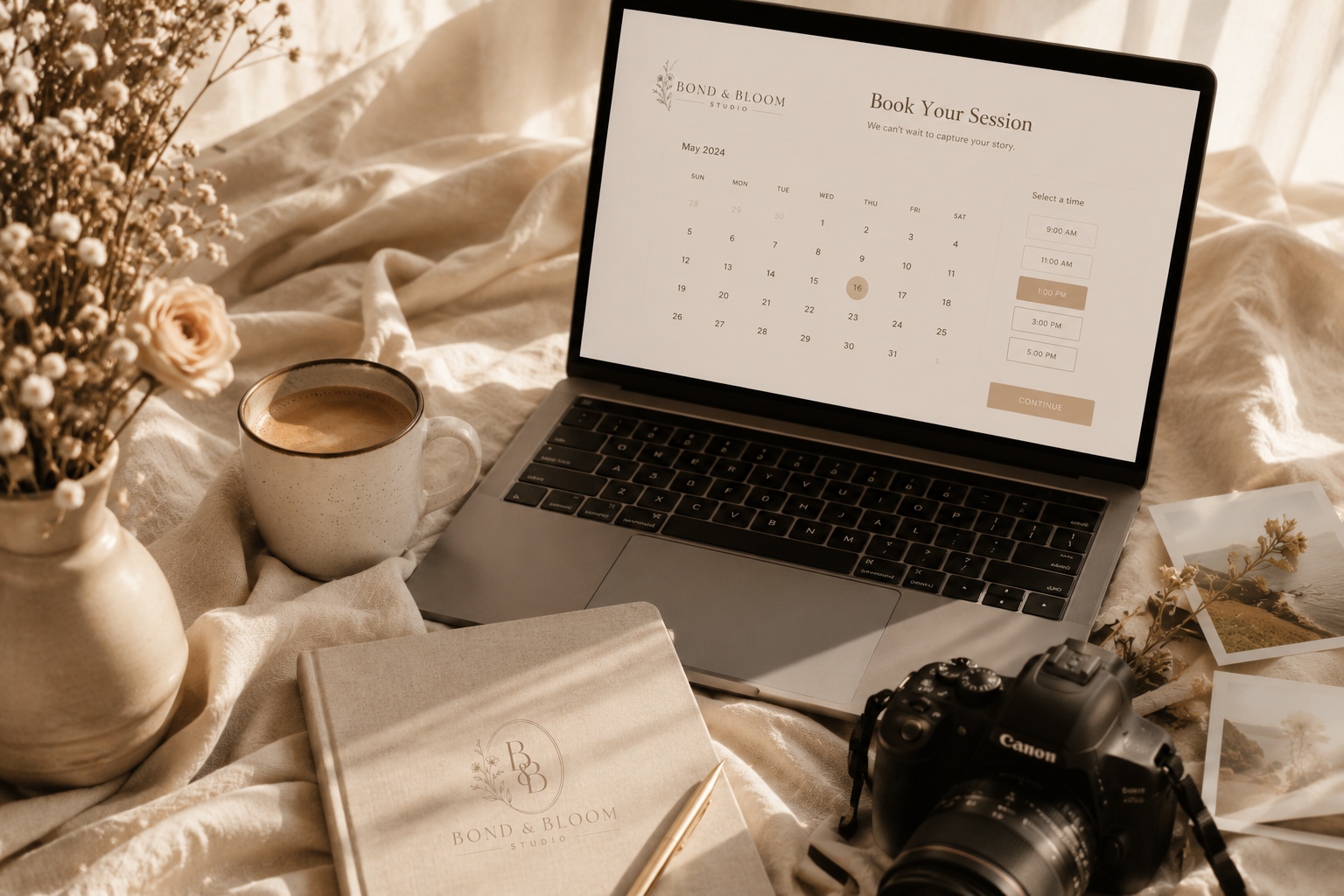 A workspace scene with a laptop showing a booking calendar for Bond & Bloom Studio, a cup of coffee, a vase with flowers, a notebook, a pen, a camera, and photographs on a bed or soft surface.