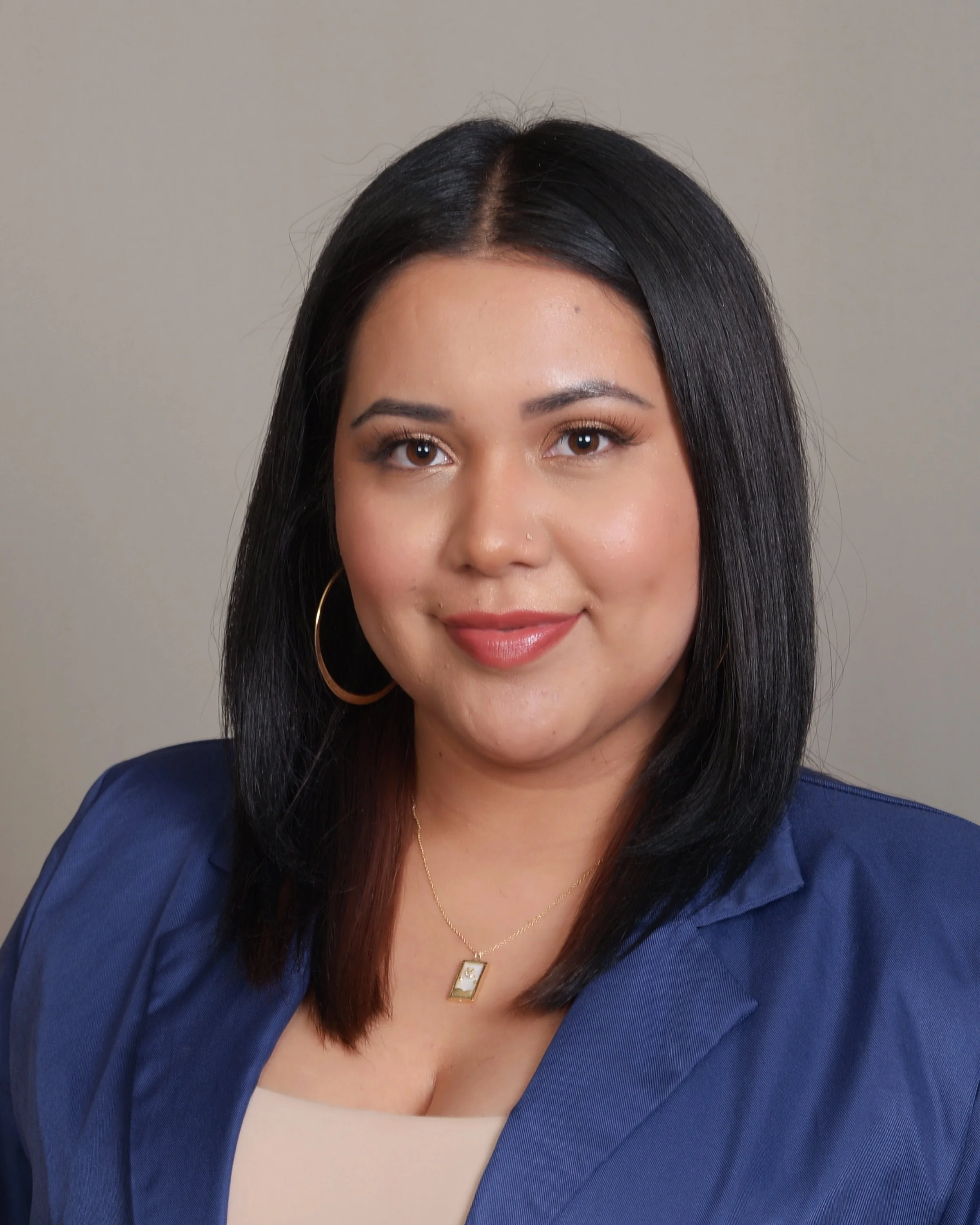 A woman with shoulder-length black hair, wearing gold hoop earrings, a gold necklace with a rectangular pendant, and a blue blazer, smiling, against a plain gray background.