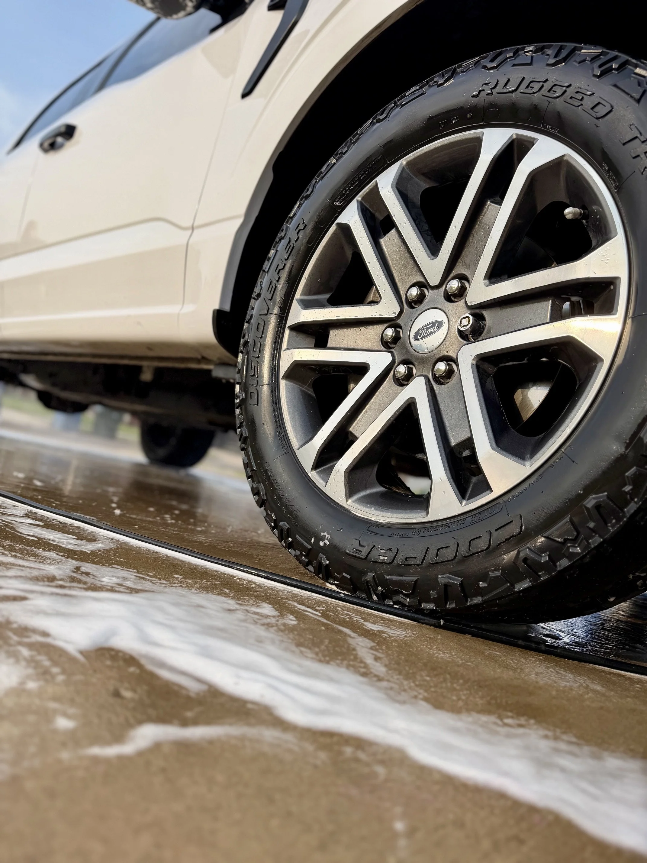 Close-up of a car tire with a Ford logo on the rim, wet from water, near the edge of a wet surface with foam or soap residue.