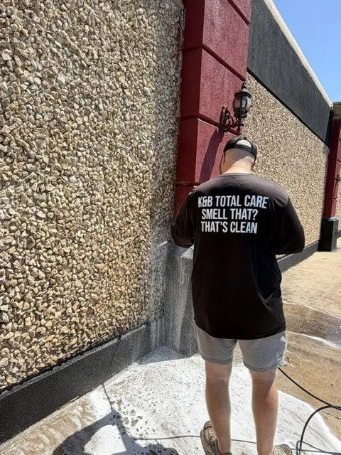 Person wearing a black sweatshirt with white text reading 'K&B TOTAL CARE, SMELL THAT? THAT'S CLEAN' power washing a wall outside a building with textured walls and red columns.