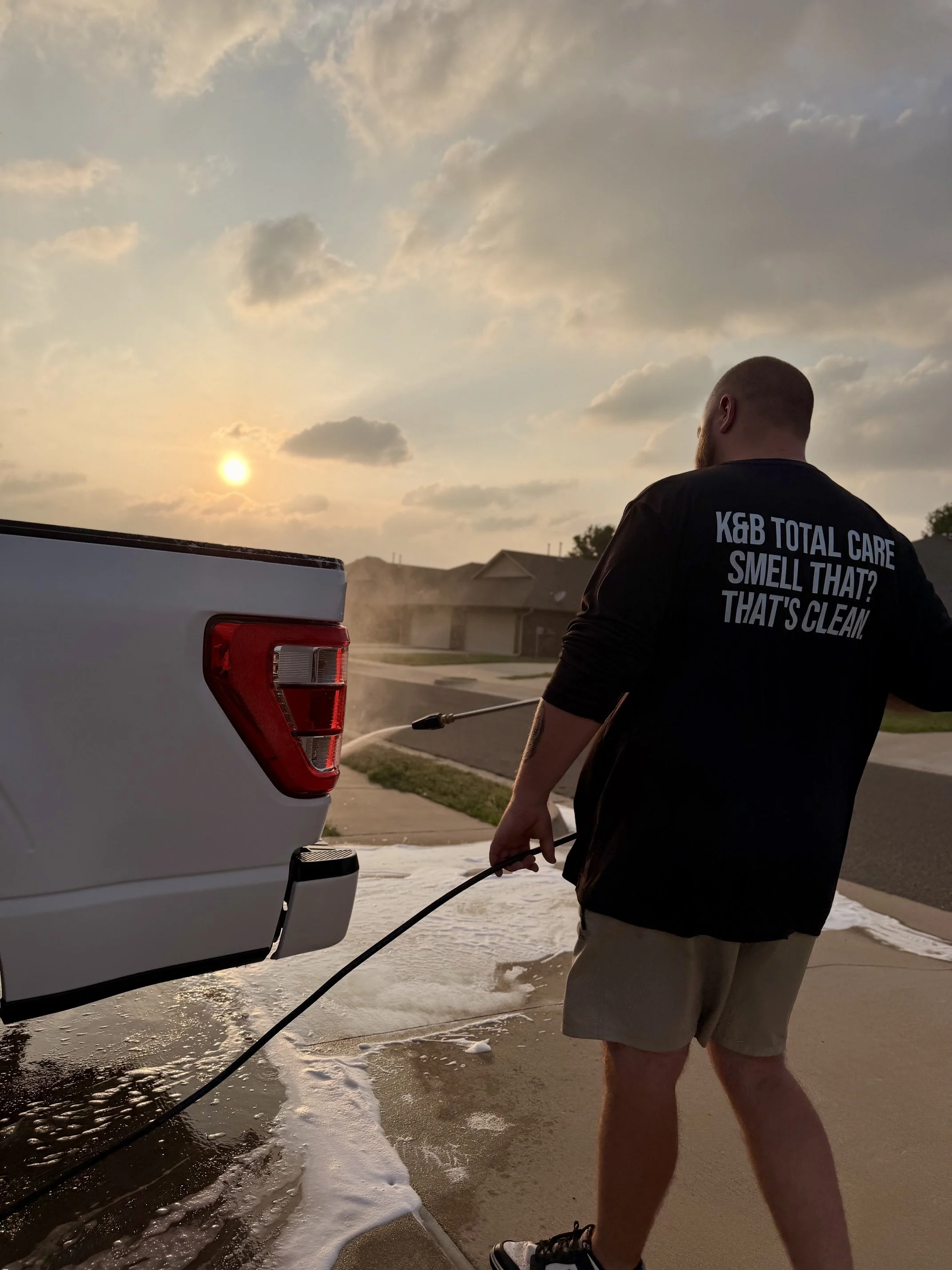 A man with a beard wearing a black T-shirt and beige shorts is pressure washing a white truck. The back of the man's T-shirt reads: 'K&B Total Care Smell That? That's Clean.' 