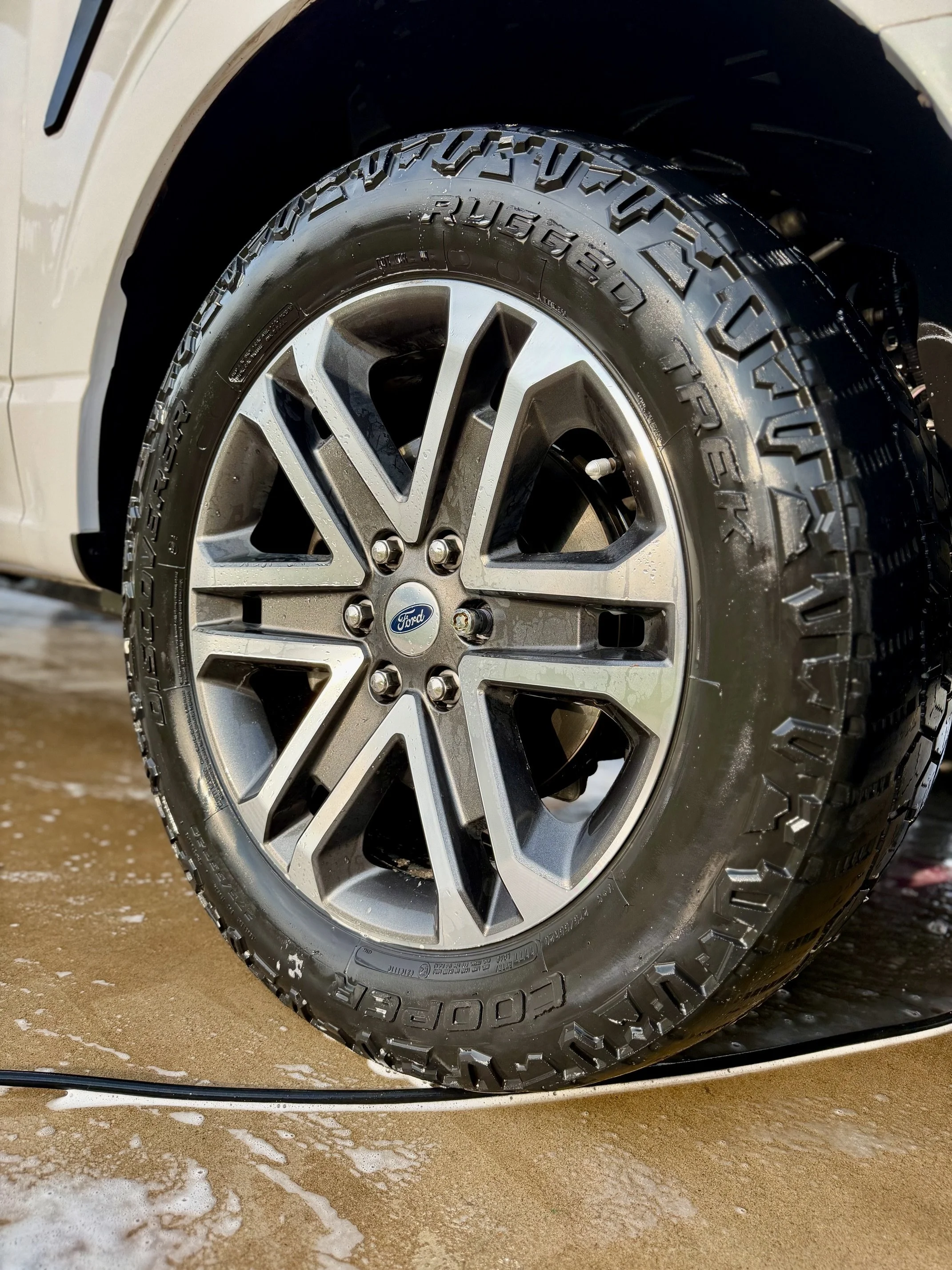 Close-up of a glossy and clean vehicle tire, mounted on a silver alloy wheel with a Ford logo at the center, on a wet surface with soap suds around.