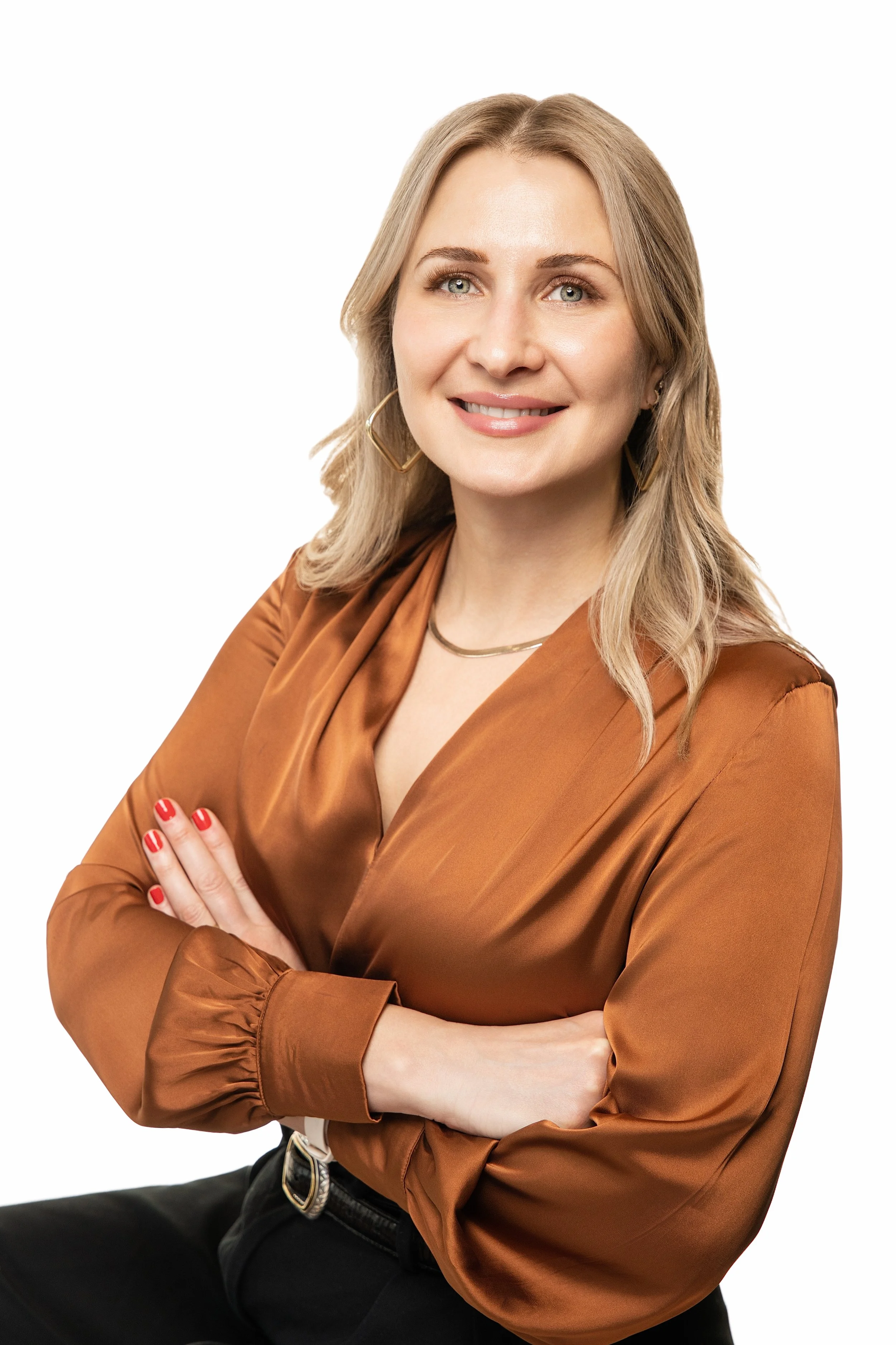 A woman with blonde hair, wearing a brown satin blouse, black pants, and gold jewelry, with her arms crossed, smiling in front of a white background.