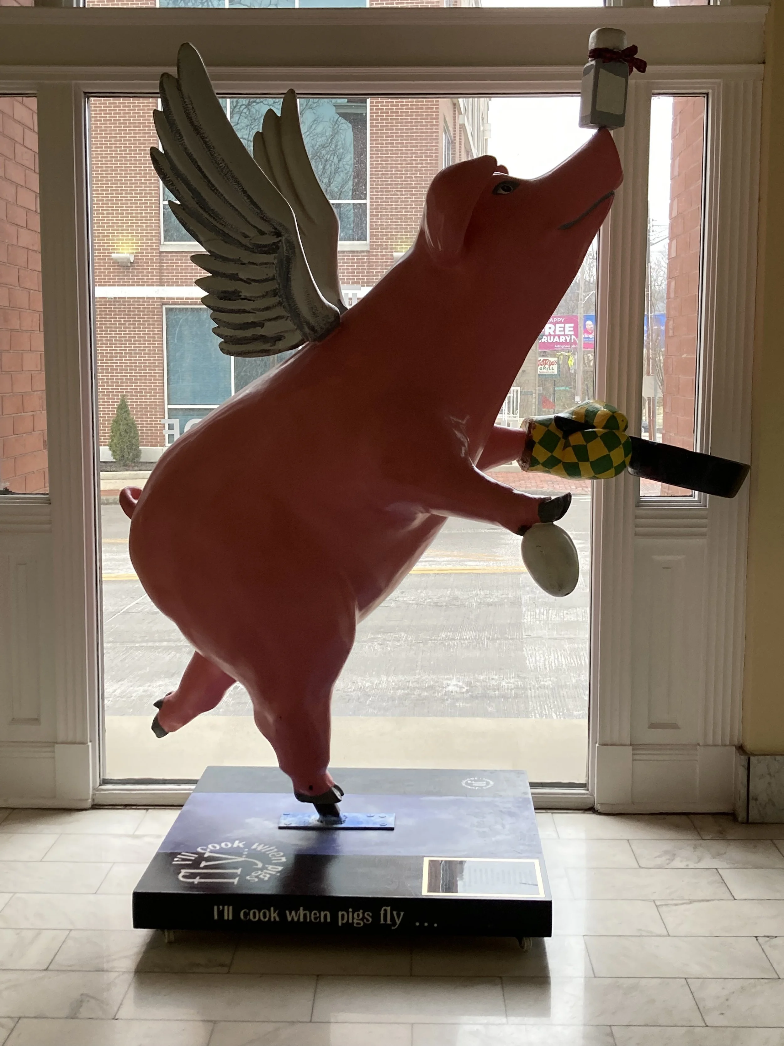 A pig-shaped sculpture with wings, standing on its hind legs inside a building near a window. The sculpture is positioned as if it is flying or jumping. The statue has a pink body, a large snout, and is wearing gloves, with a black object resembling a hammer or mallet in one hoof. The base of the sculpture has text that reads, 'I'll cook when pigs fly...'. Outside the window, a brick building and some trees are visible.
