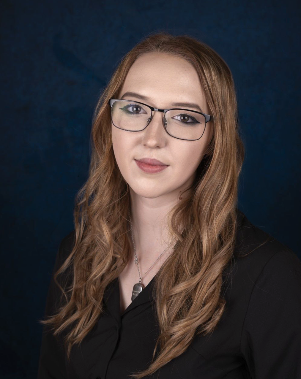 A young woman with long wavy brown hair, wearing glasses and a black top, smiling slightly against a dark blue background.