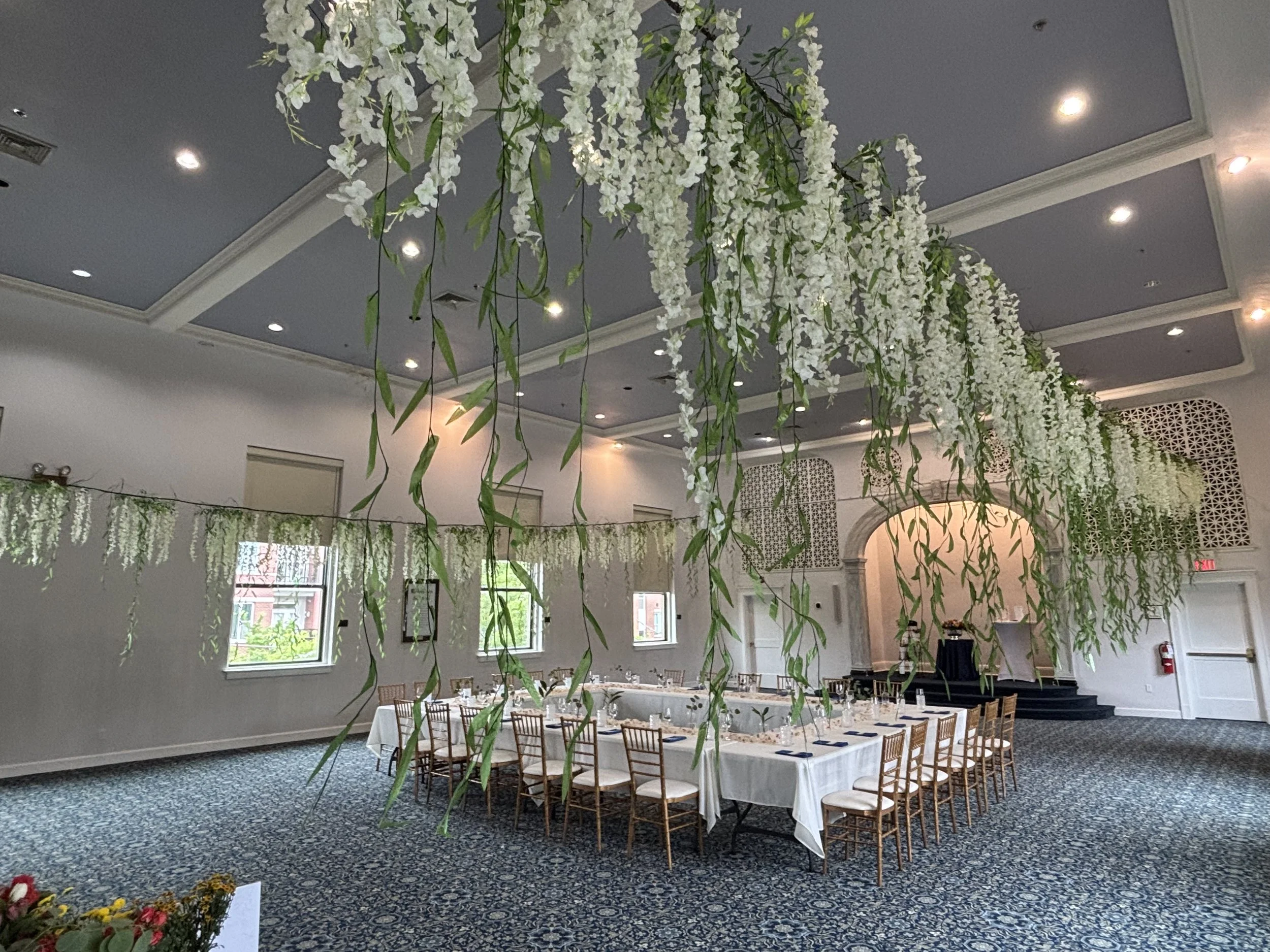 Elegant event space decorated with long table set for a formal gathering, overhead floral arrangements with cascading white flowers and green leaves, multiple windows letting in natural light, and patterned carpeted floor.