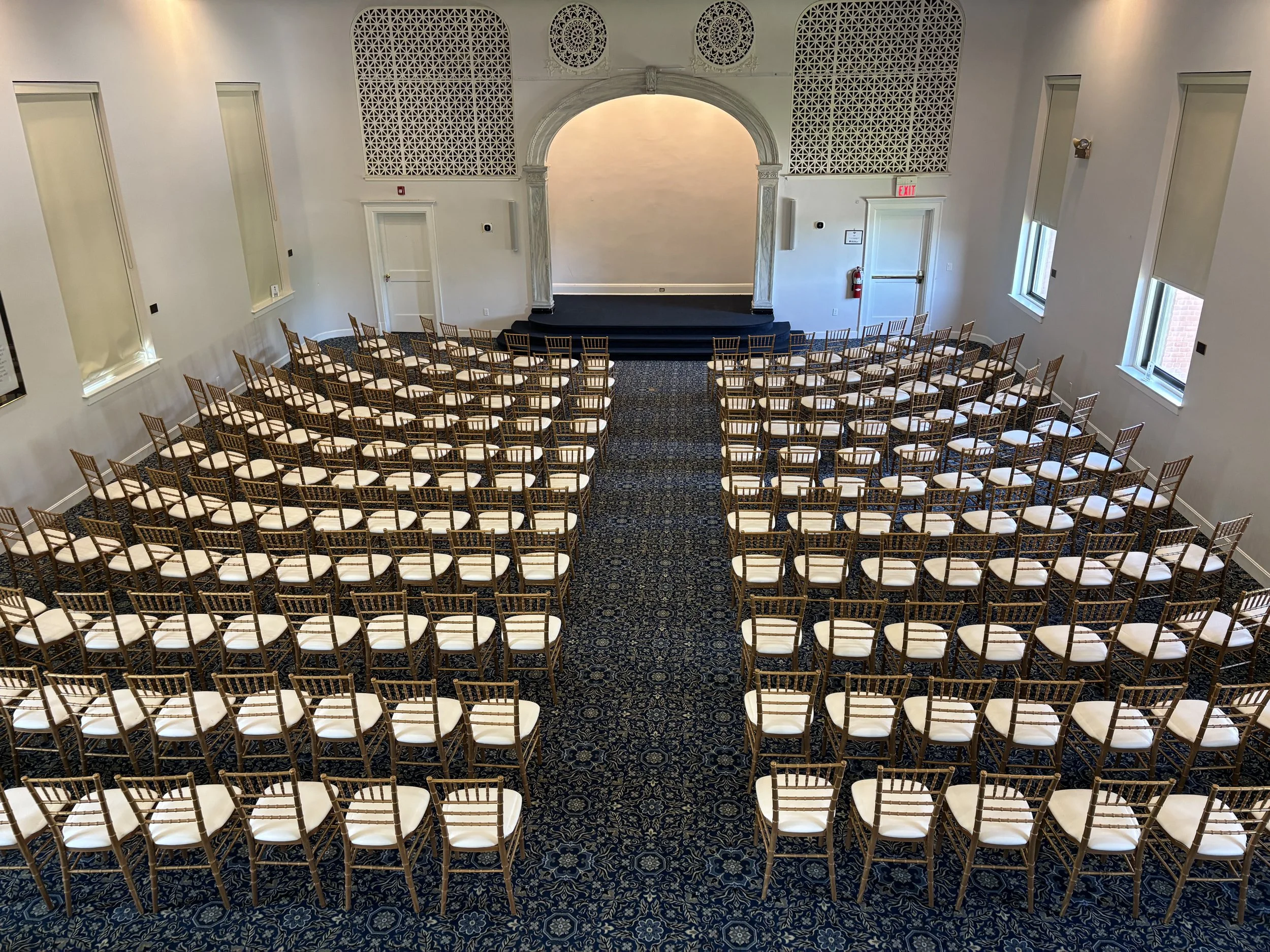 Empty auditorium with rows of white chairs with wooden backs facing a small stage with an arched backdrop in a room with high windows and decorative wall panels.