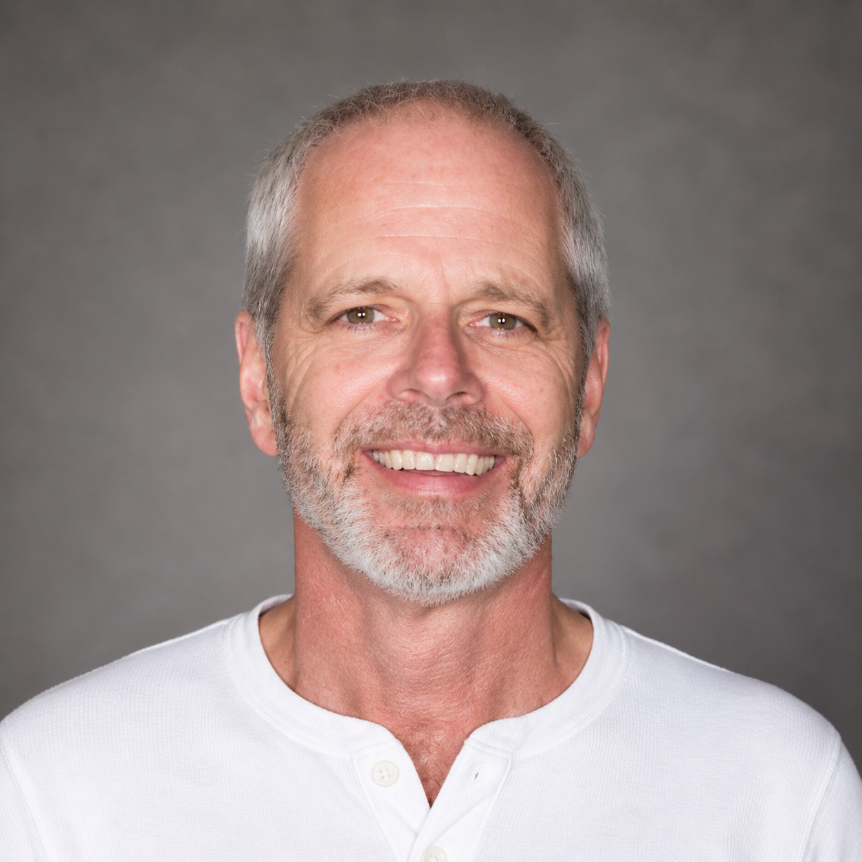 Headshot of a smiling man with gray hair, a beard, and mustache, wearing a white shirt against a gray background.
