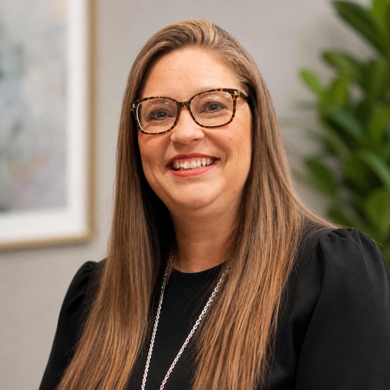A smiling woman with long brown hair and glasses, wearing a black top, in an indoor setting with a framed picture and a green plant in the background.