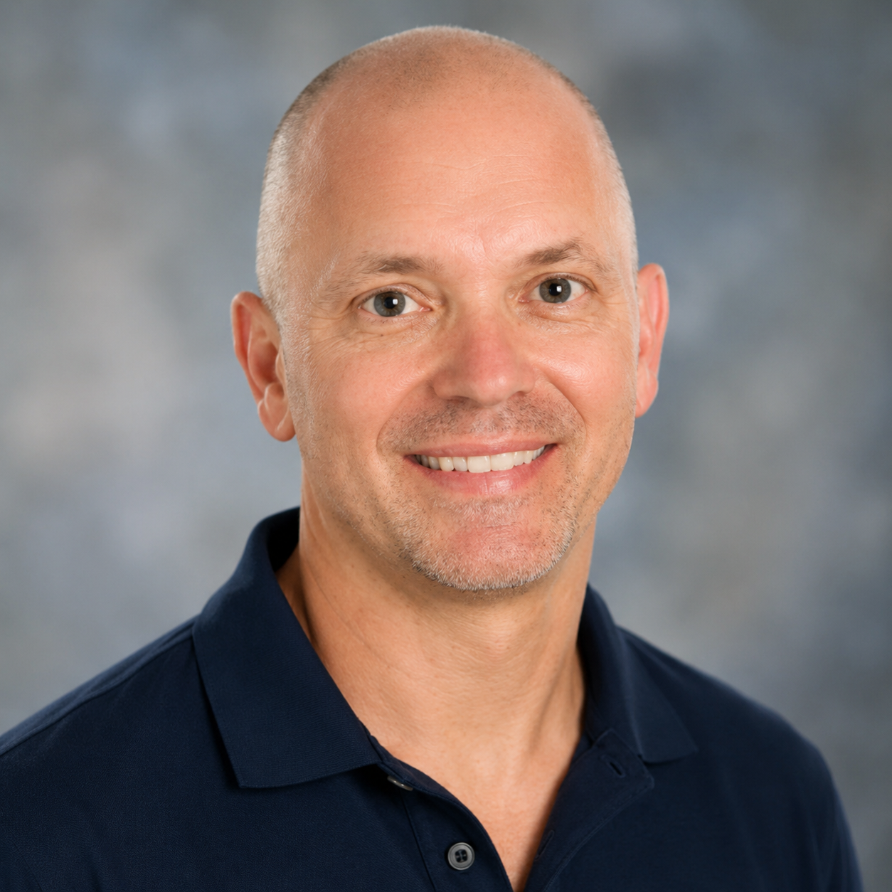Headshot of a smiling middle-aged man with a shaved head, light facial hair, wearing a navy blue polo shirt, against a neutral background.