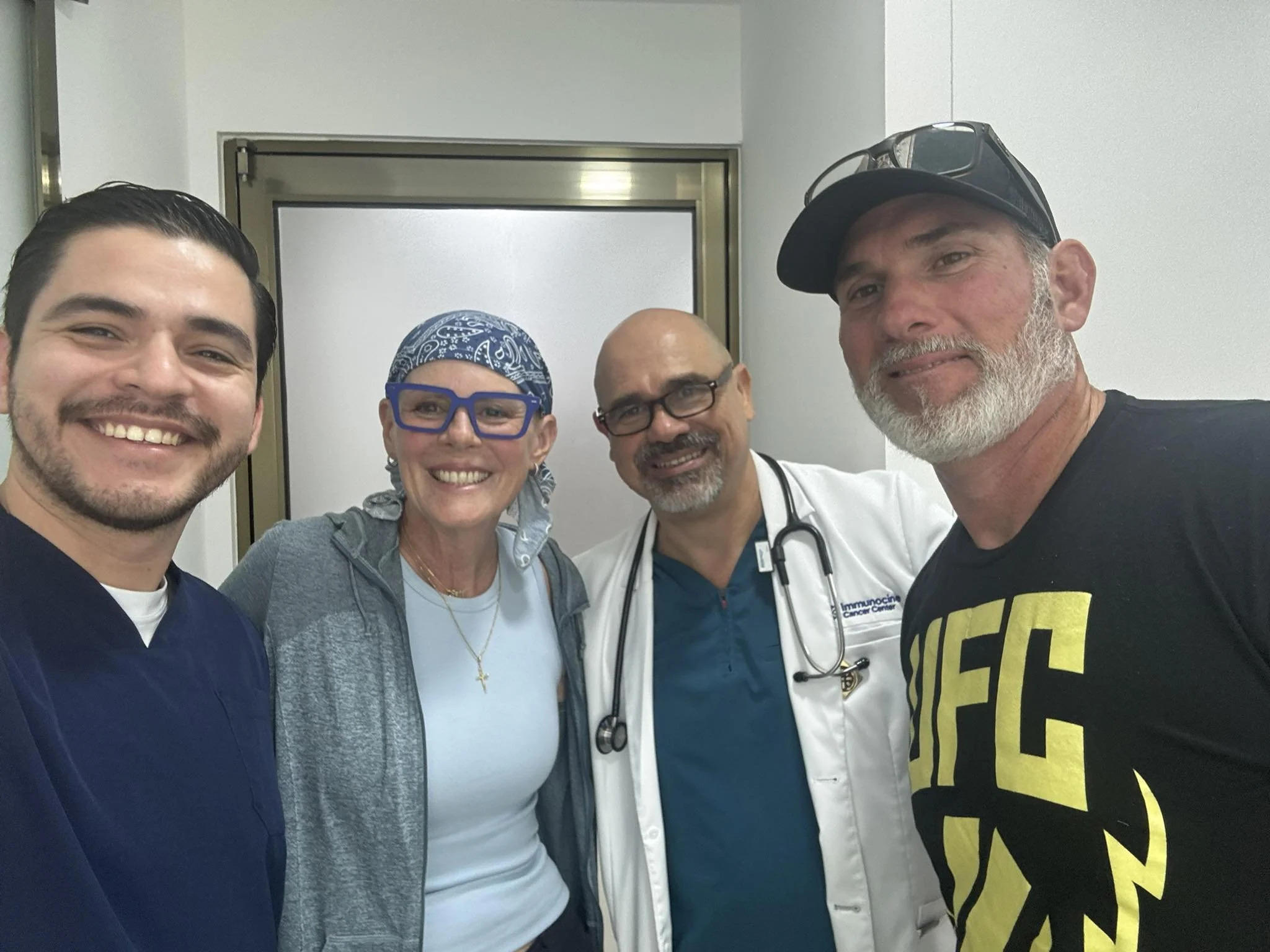 A group of four people, including a medical professional wearing scrubs and stethoscope, taking a selfie together in a medical setting. They are smiling, and the background shows the entrance to an elevator or door in a hospital or clinic.