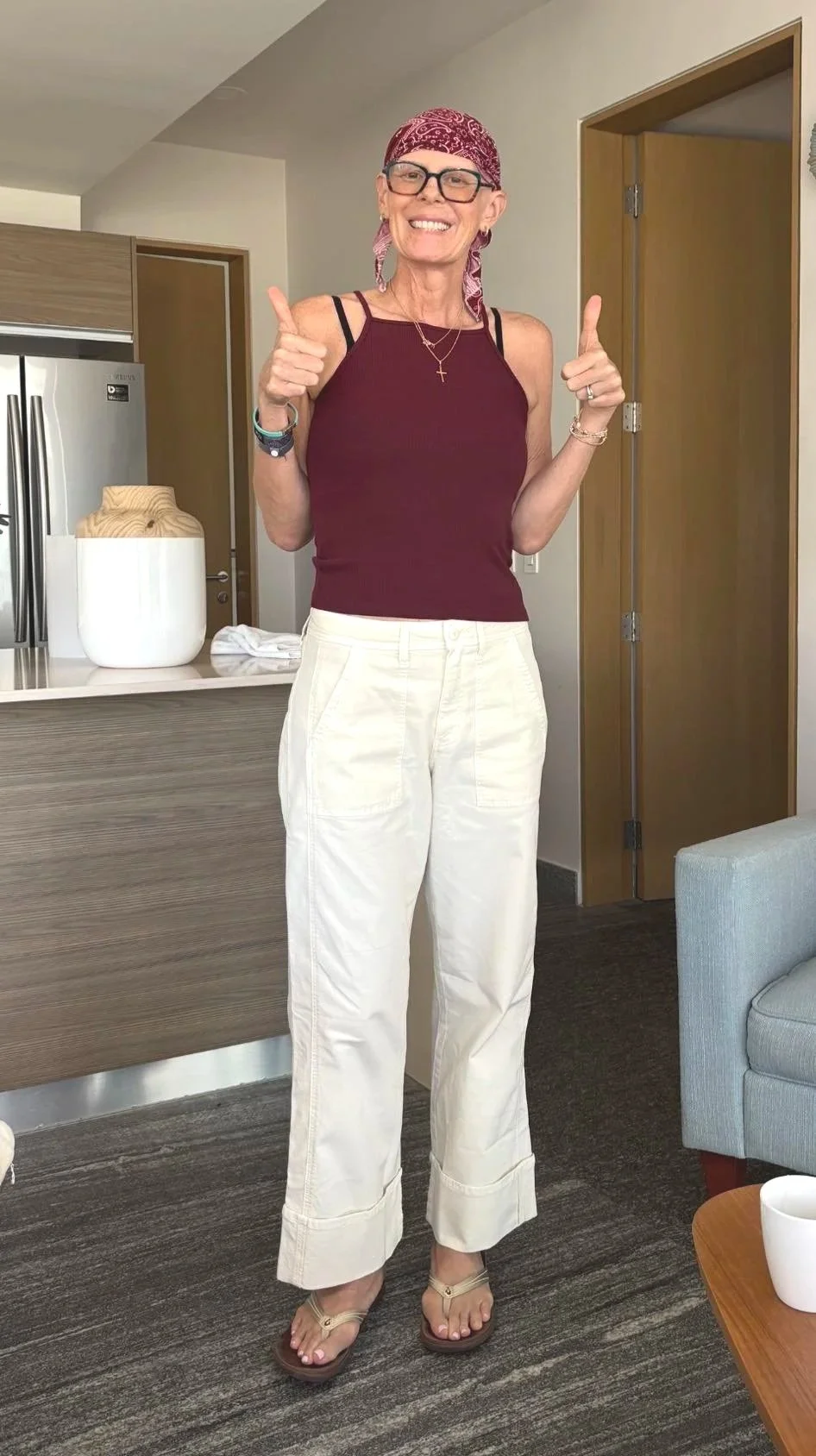 A smiling woman wearing a red bandana, glasses, a maroon tank top, and white wide-leg pants standing indoors. She is giving thumbs up with both hands.
