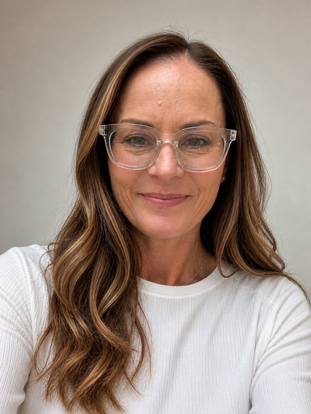 A smiling woman with long wavy brown hair, wearing clear glasses and a white ribbed shirt.